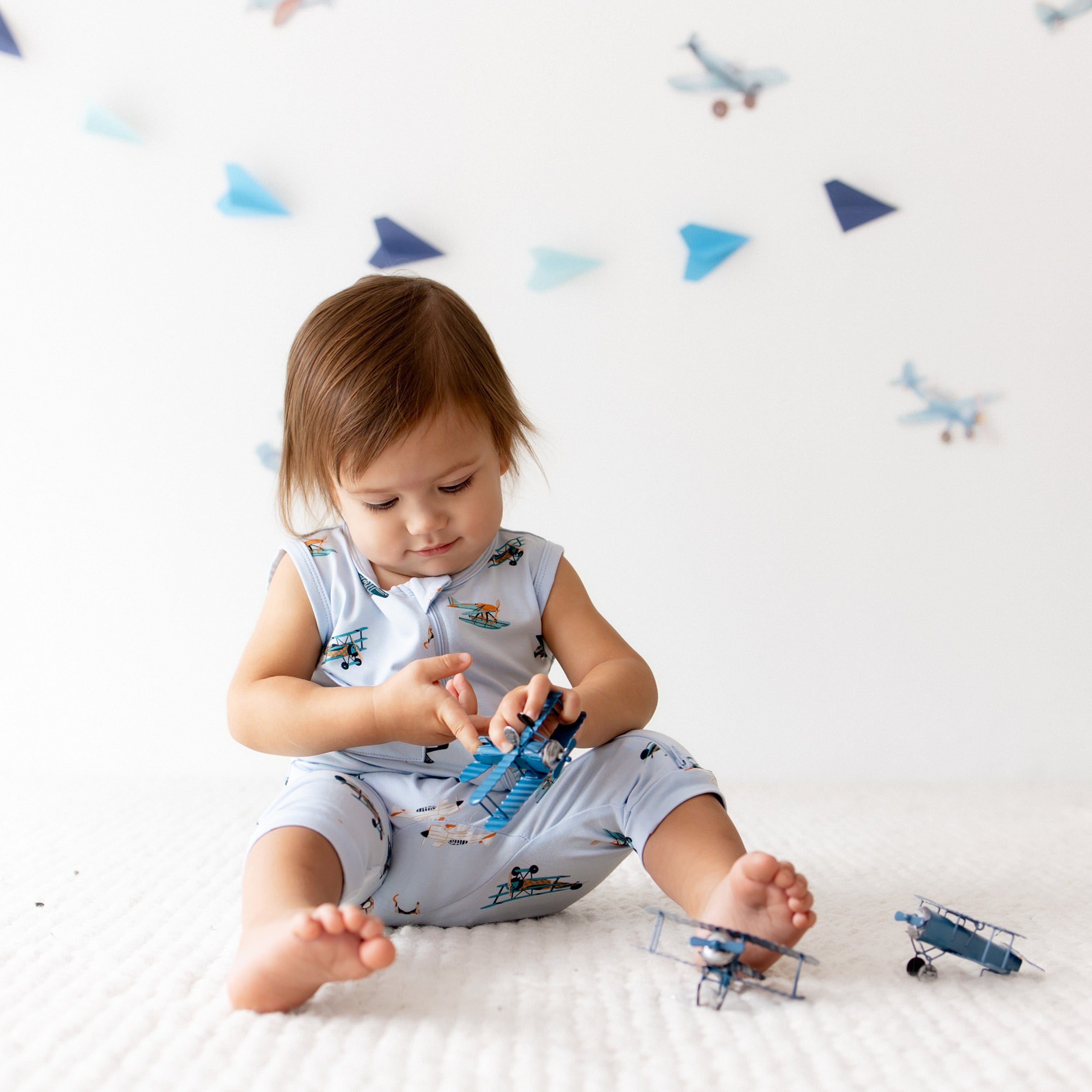 Young toddler sitting on the floor wearing the Zippered Sleeveless Romper in Take Off playing with toy planes