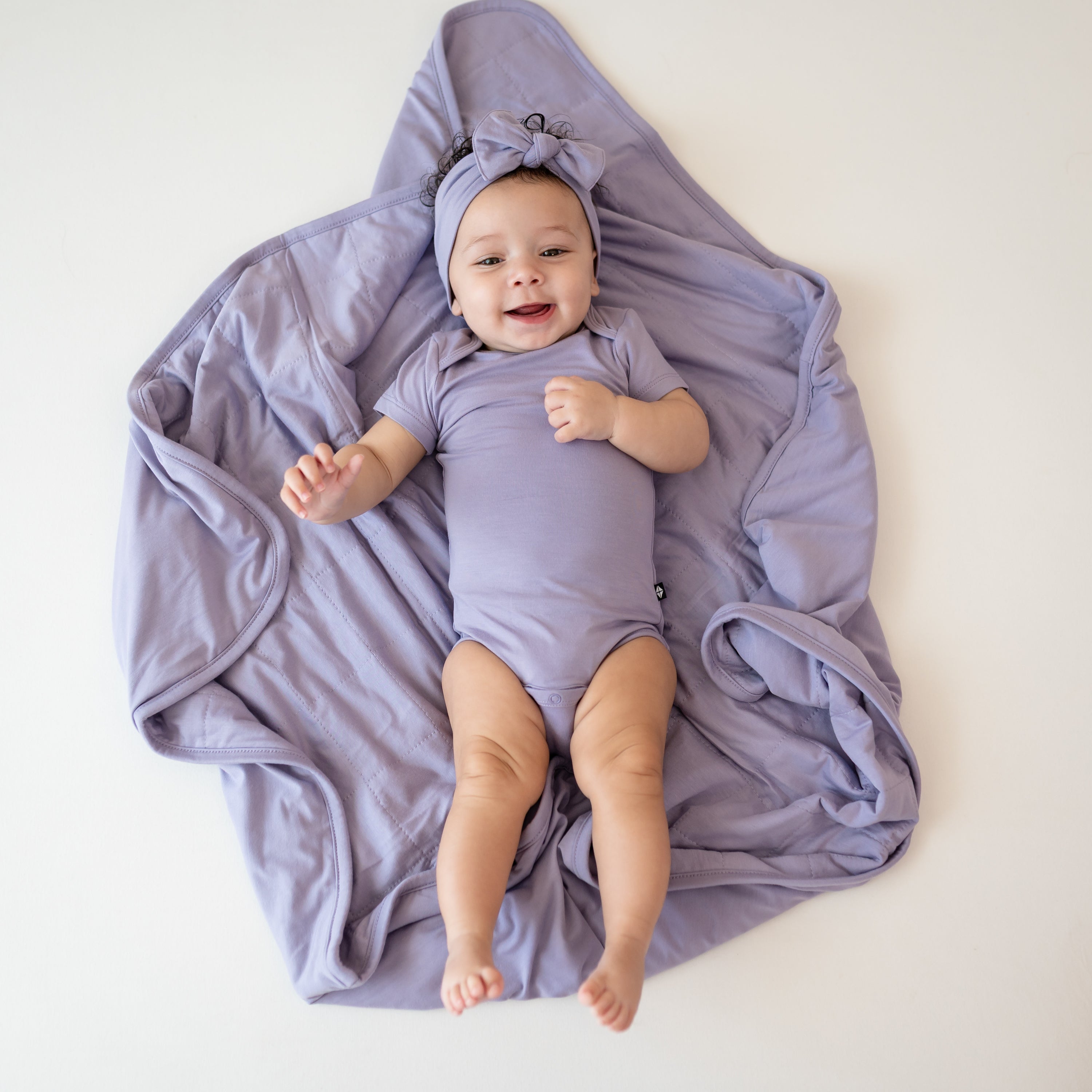 Baby laying on Baby Blanket in Taro 1.0 wearing bodysuit and bow in taro
