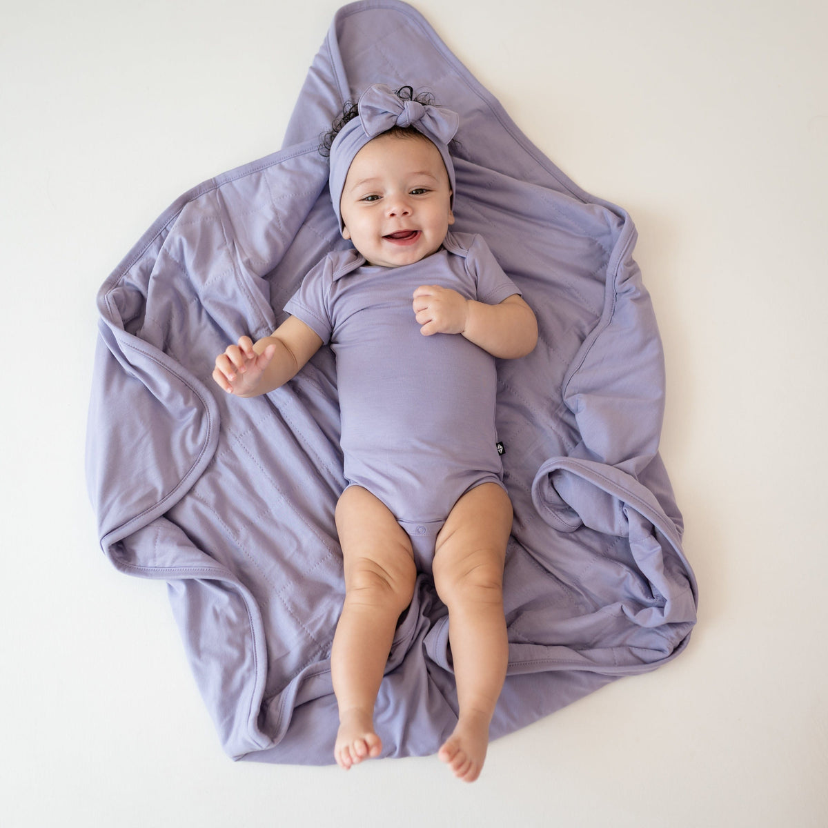 Baby laying on Baby Blanket in Taro 1.0 wearing bodysuit and bow in taro