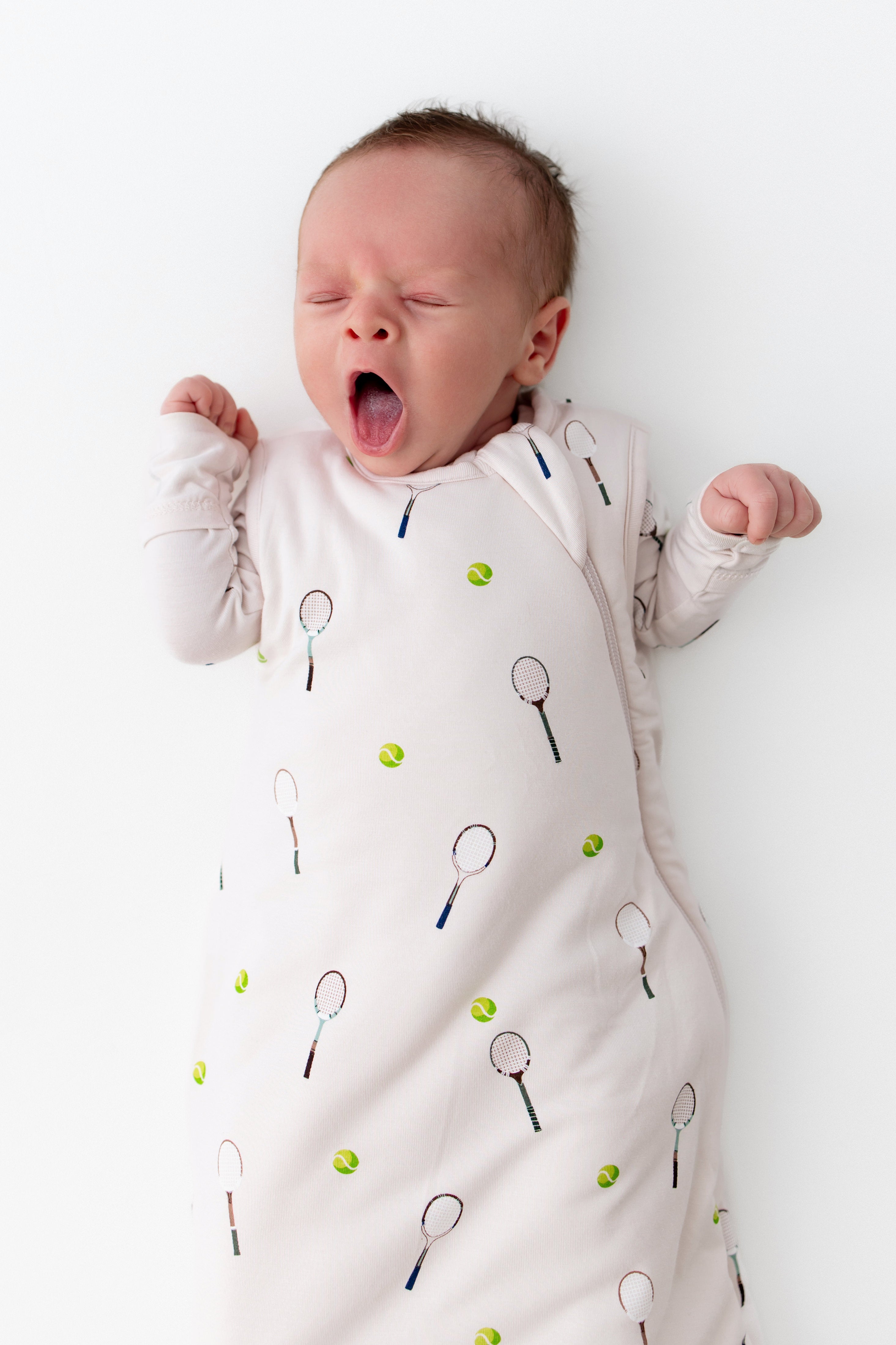 Newborn baby wearing a sleep sack with tennis racket and ball pattern on a white background
