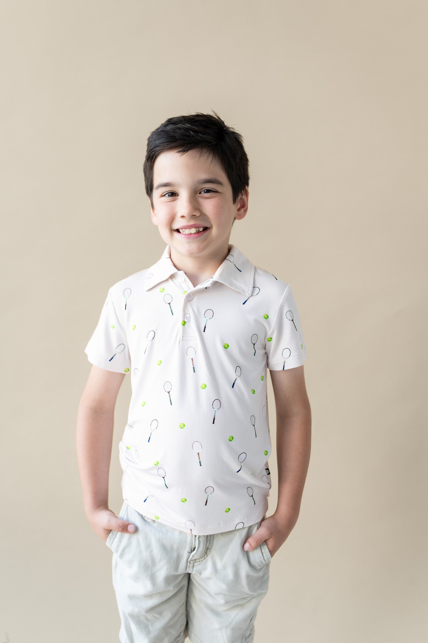 Young boy wearing a white polo shirt with a tennis-themed pattern on a beige background