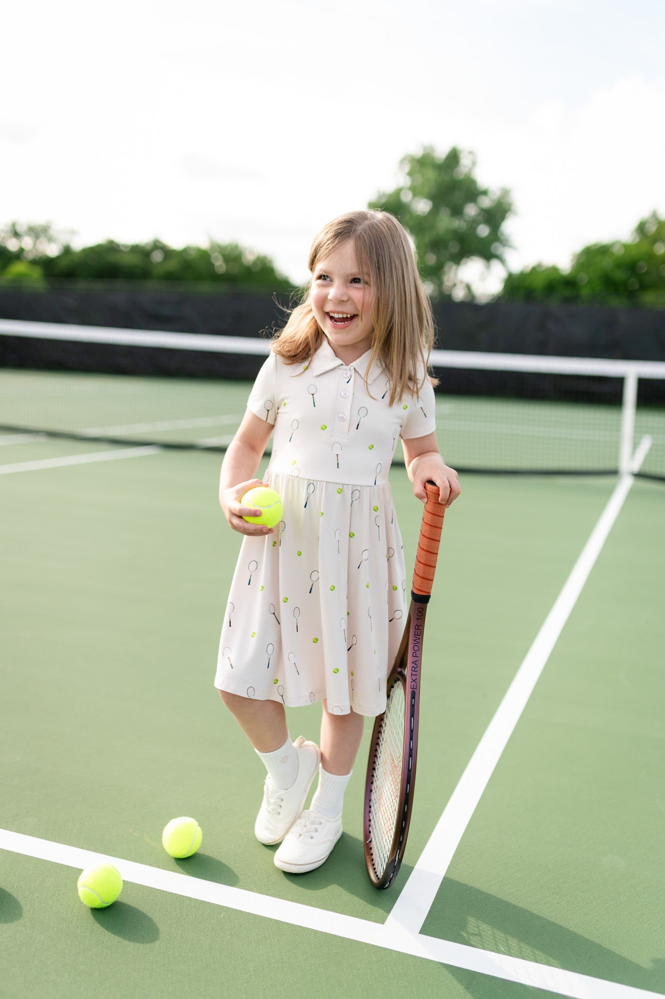Young girl in a polo dress with tennis pattern on a tennis court holding a racket and a ball