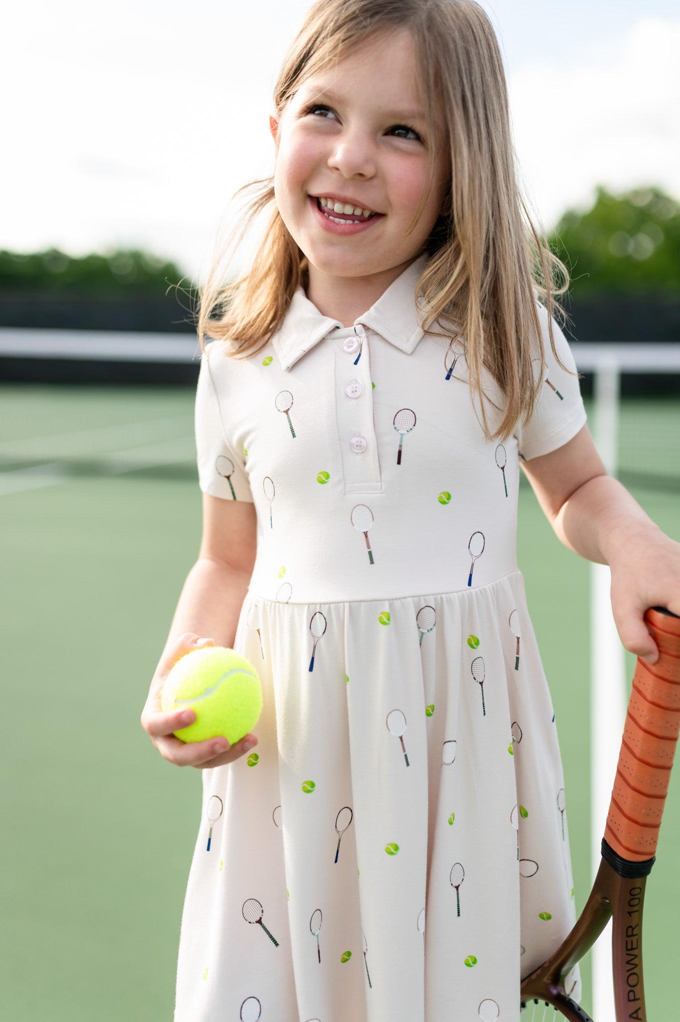 Young girl in a tennis-themed dress holding a tennis ball and racket on a tennis court.