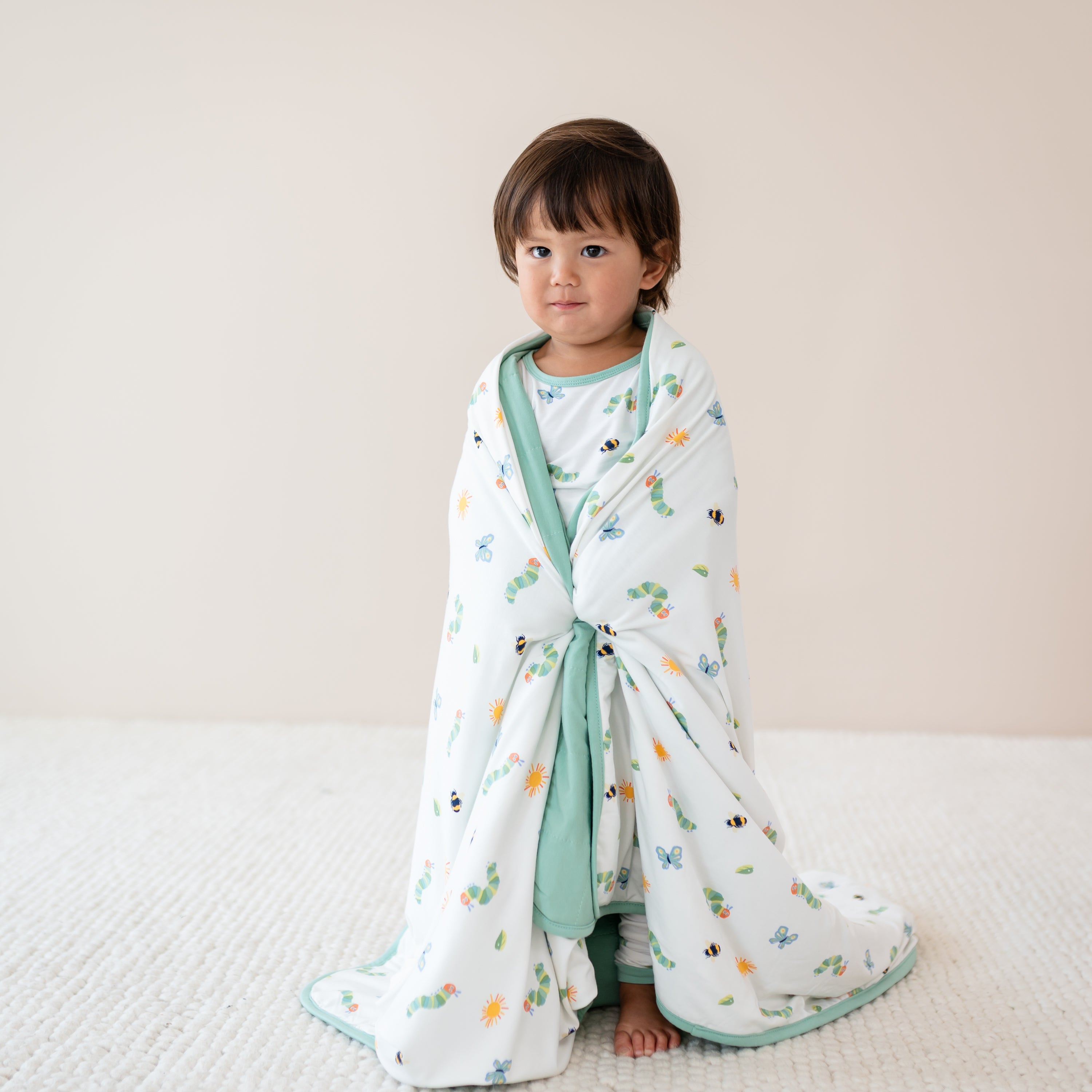 Child wrapped in a blanket in the print The Very Hungry Caterpillar print wearing matching pajamas on a plain background