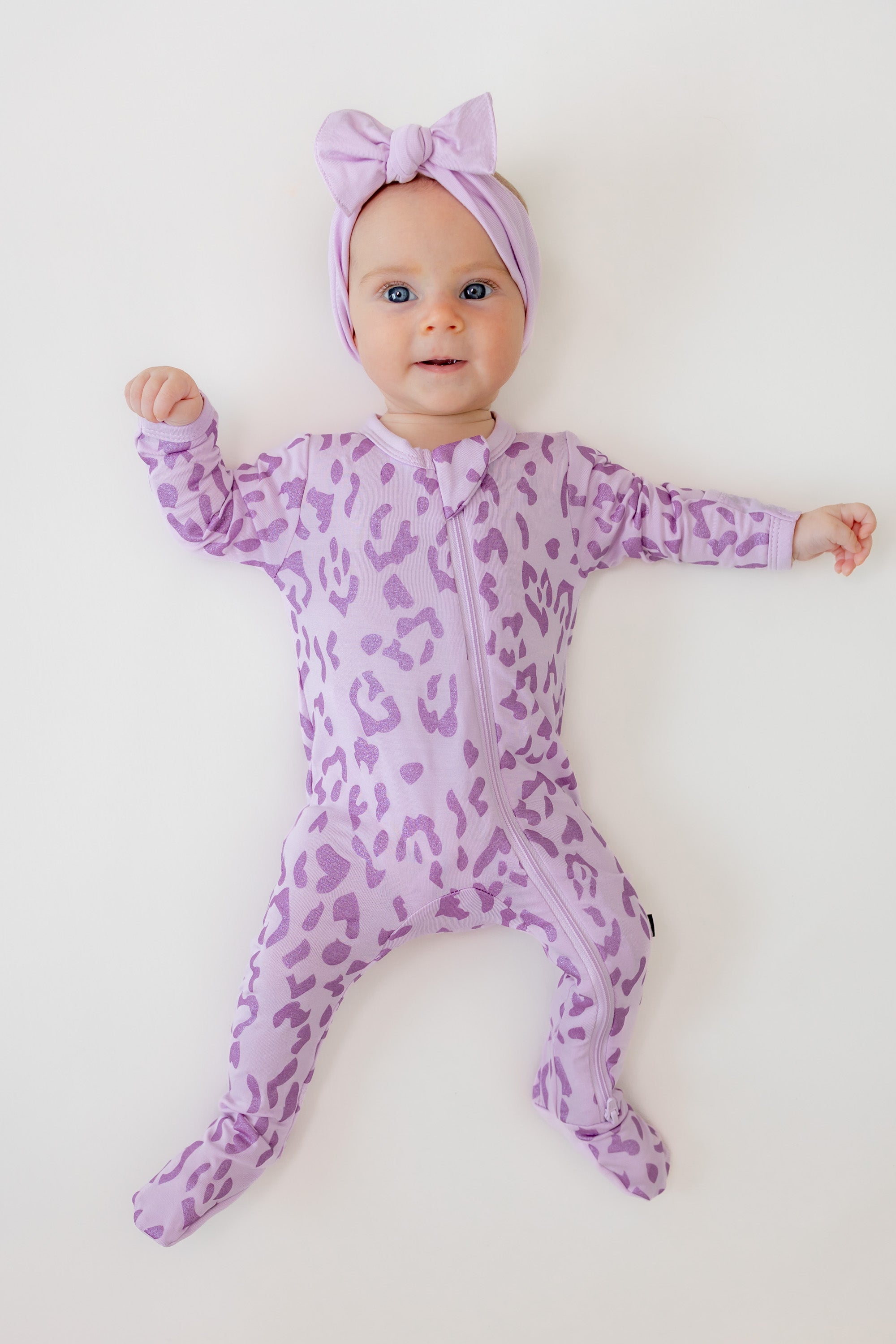 Baby wearing zippered footie in Thistle Leopard print with a thistle bow headband on a beige background