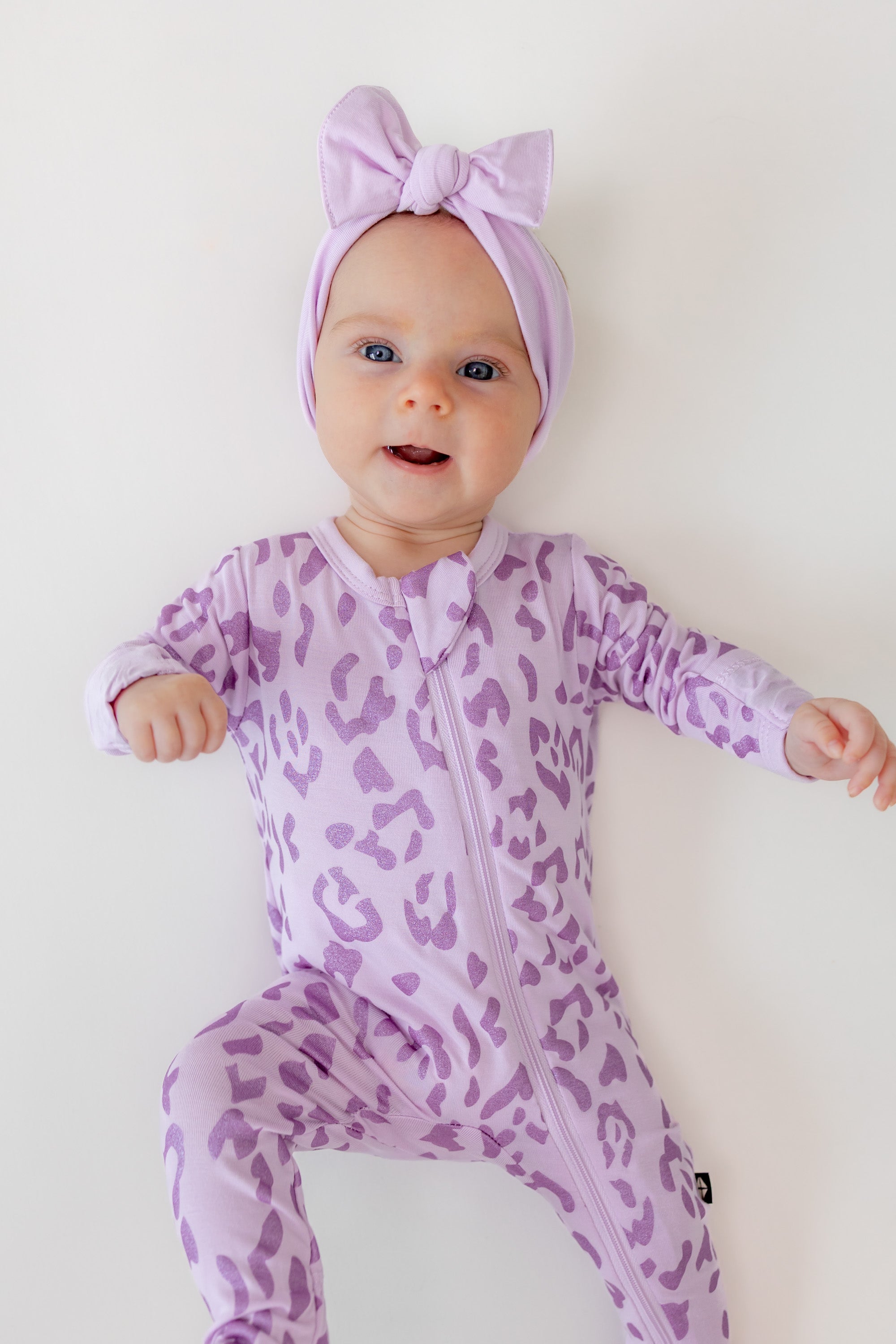 Baby wearing zippered footie in Thistle Leopard print with a thistle bow headband on a beige background