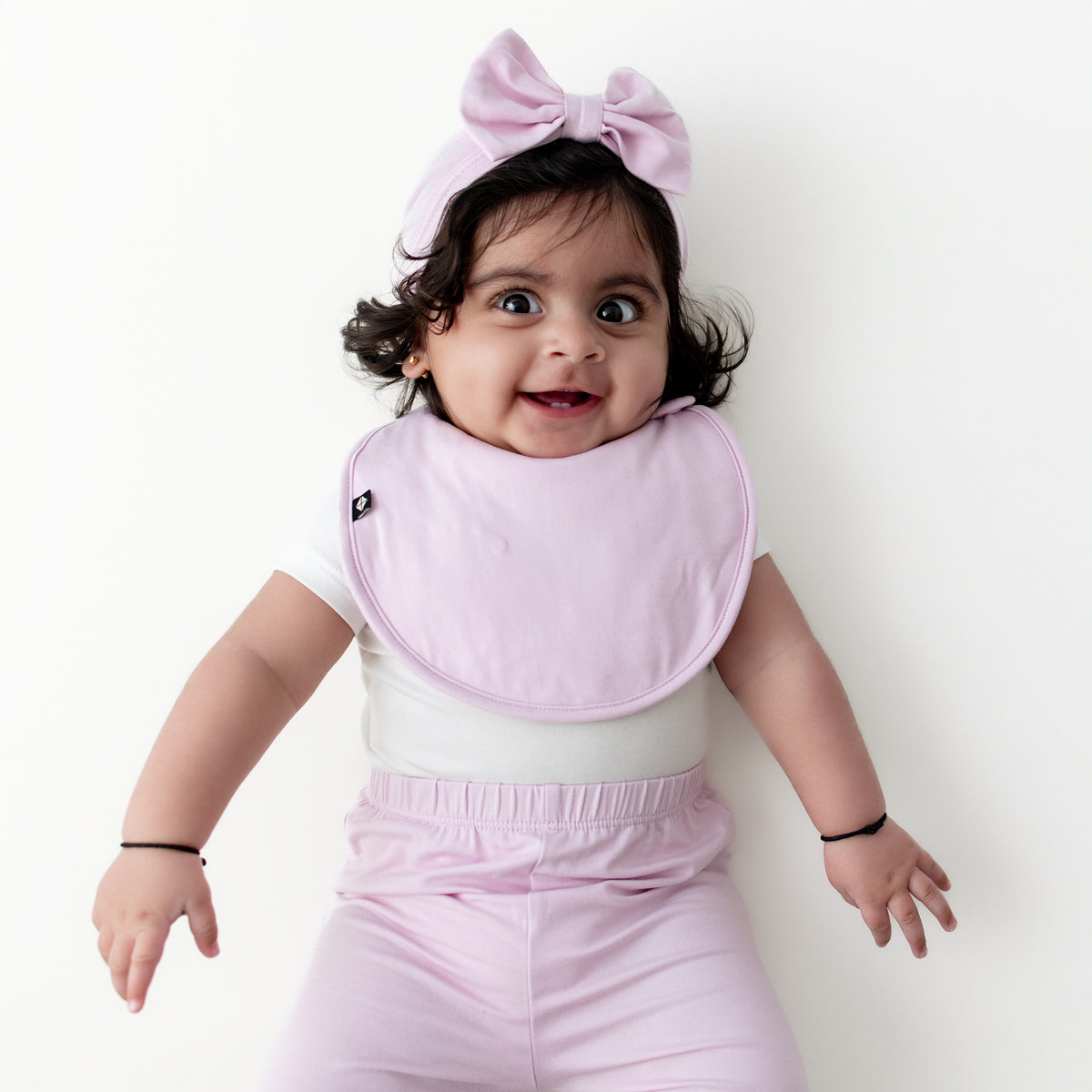Infant girl wearing the Cushy Bib in Thistle with matching bow headwrap and pants