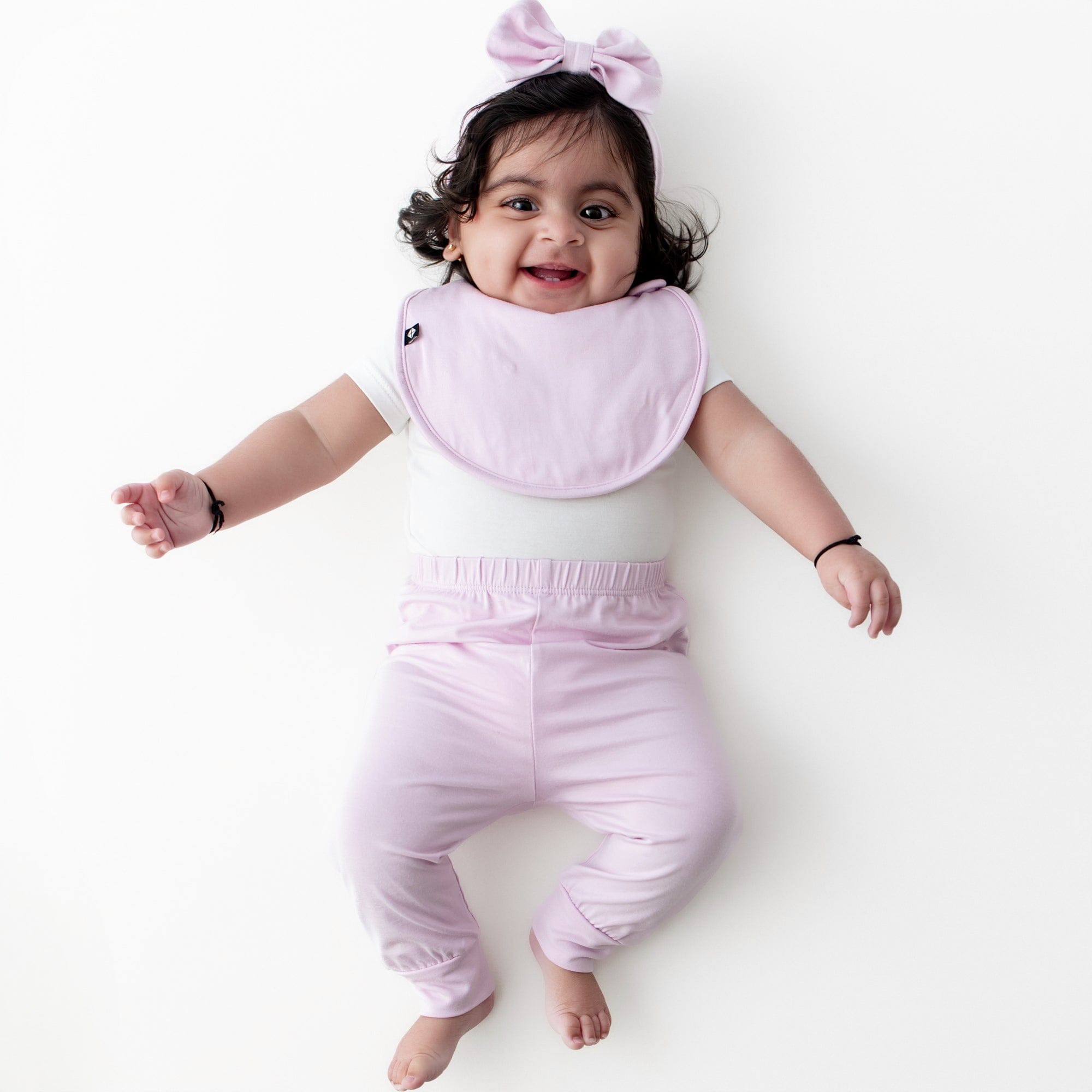 Smiling girl wearing the Cushy Bib in Thistle with matching pants and bow headwrap with a white short sleeve bodysuit