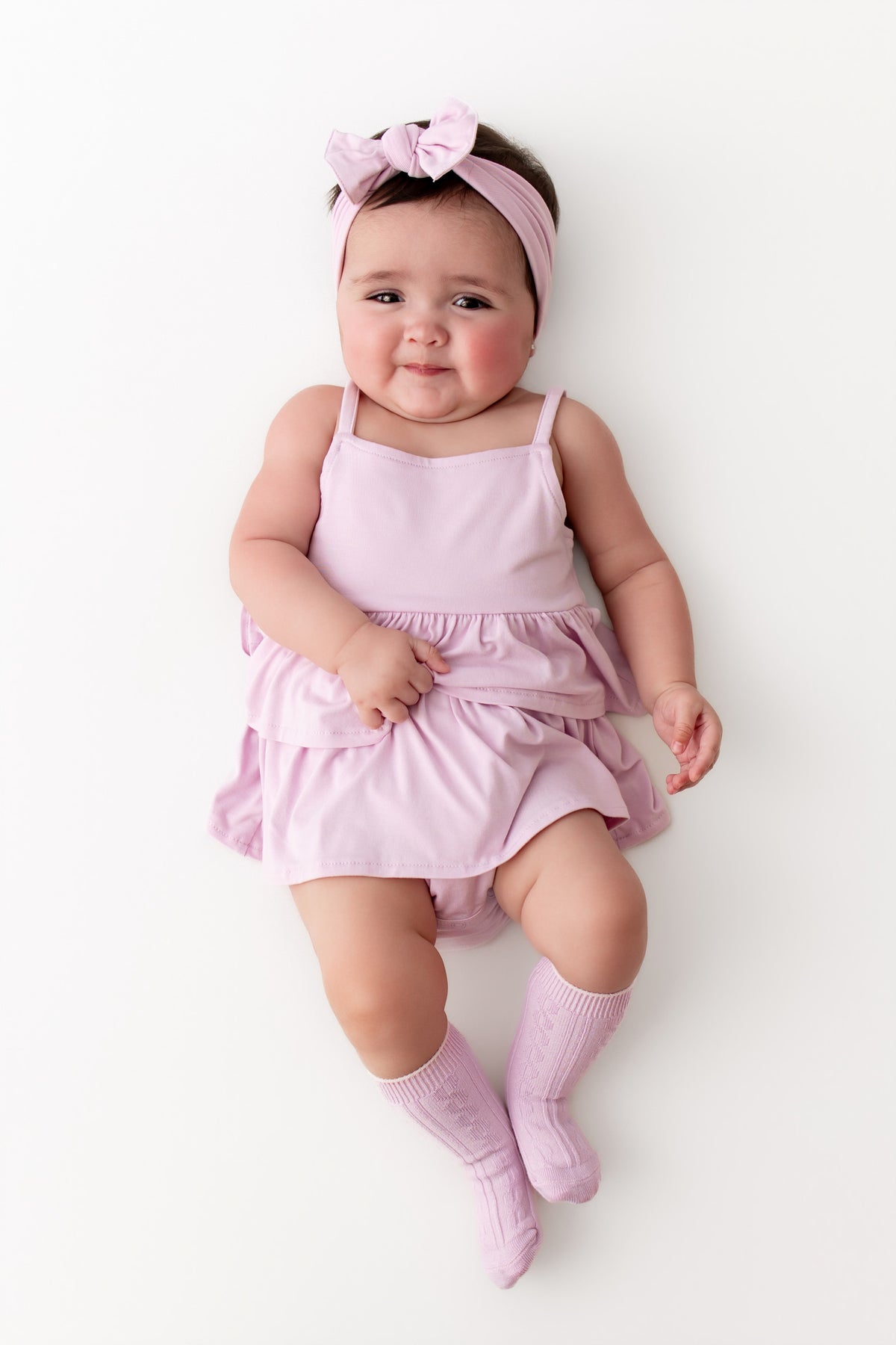 A baby lying on a white background wearing a light pink sleeveless romper dress with a ruffled skirt, a matching pink bow headband, and pink knit socks.