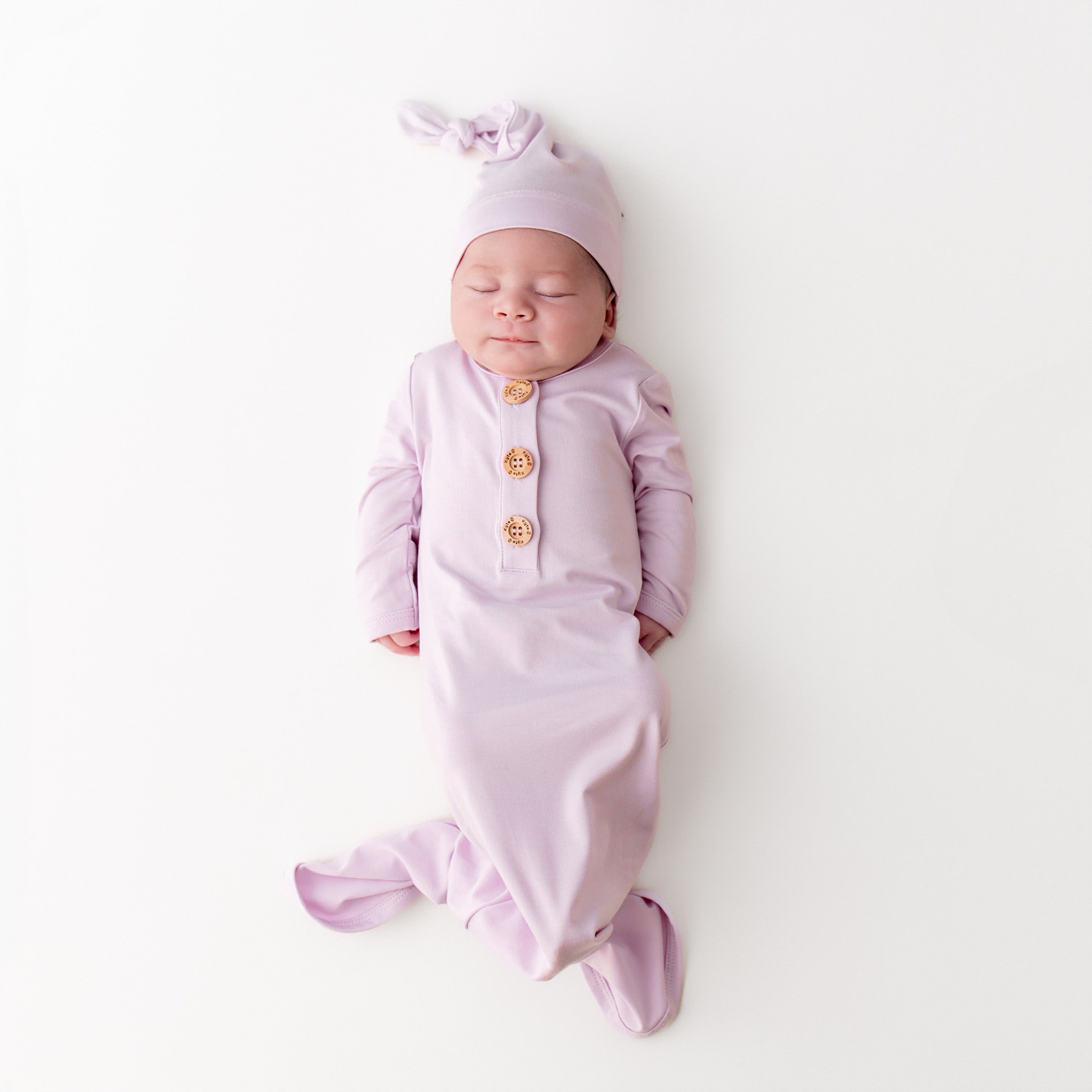 Sleeping newborn wearing the Knotted Gown with Hat Set in Thistle laying on a white surface