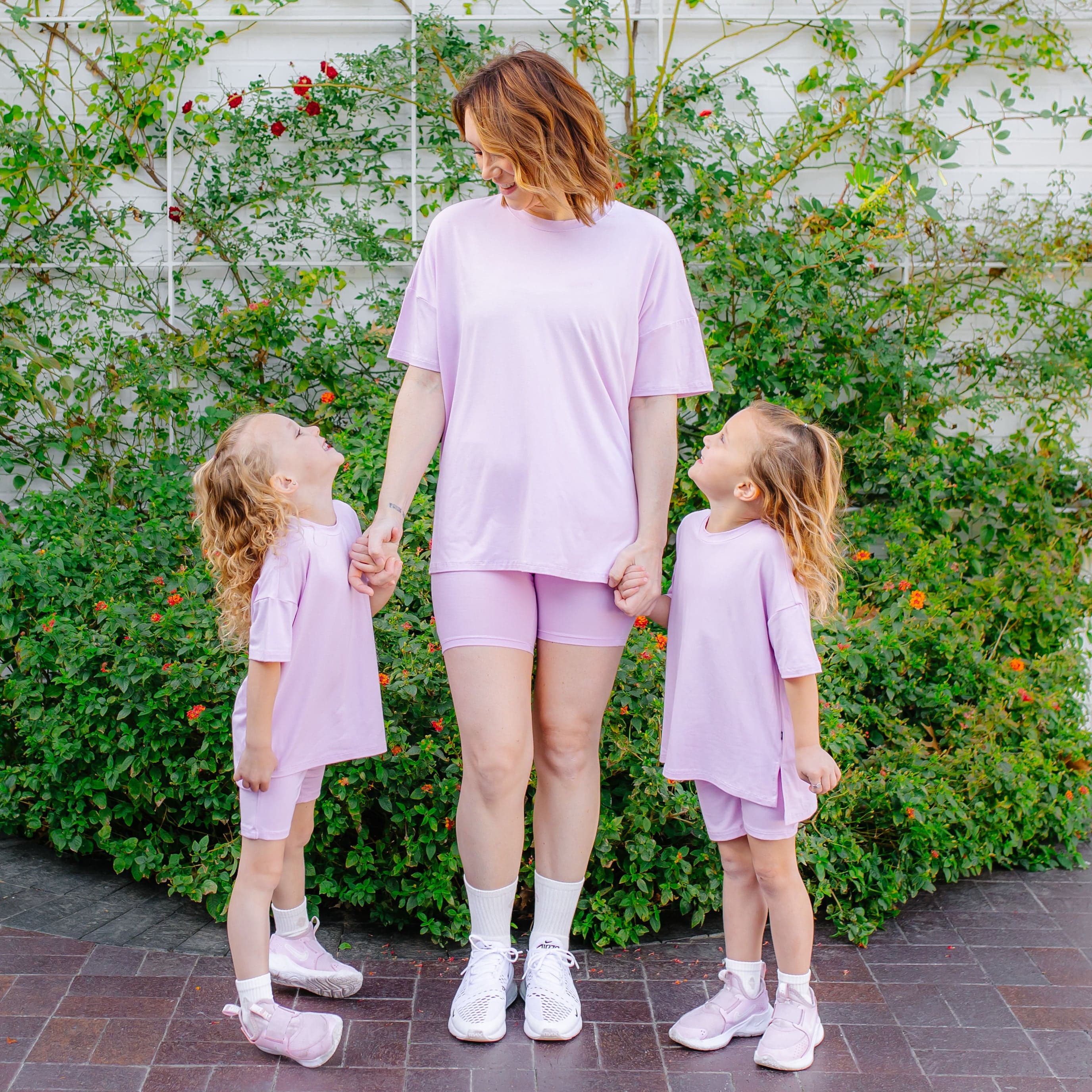 Woman and two young girls in matching lavender outfits standing outdoors with greenery in the background