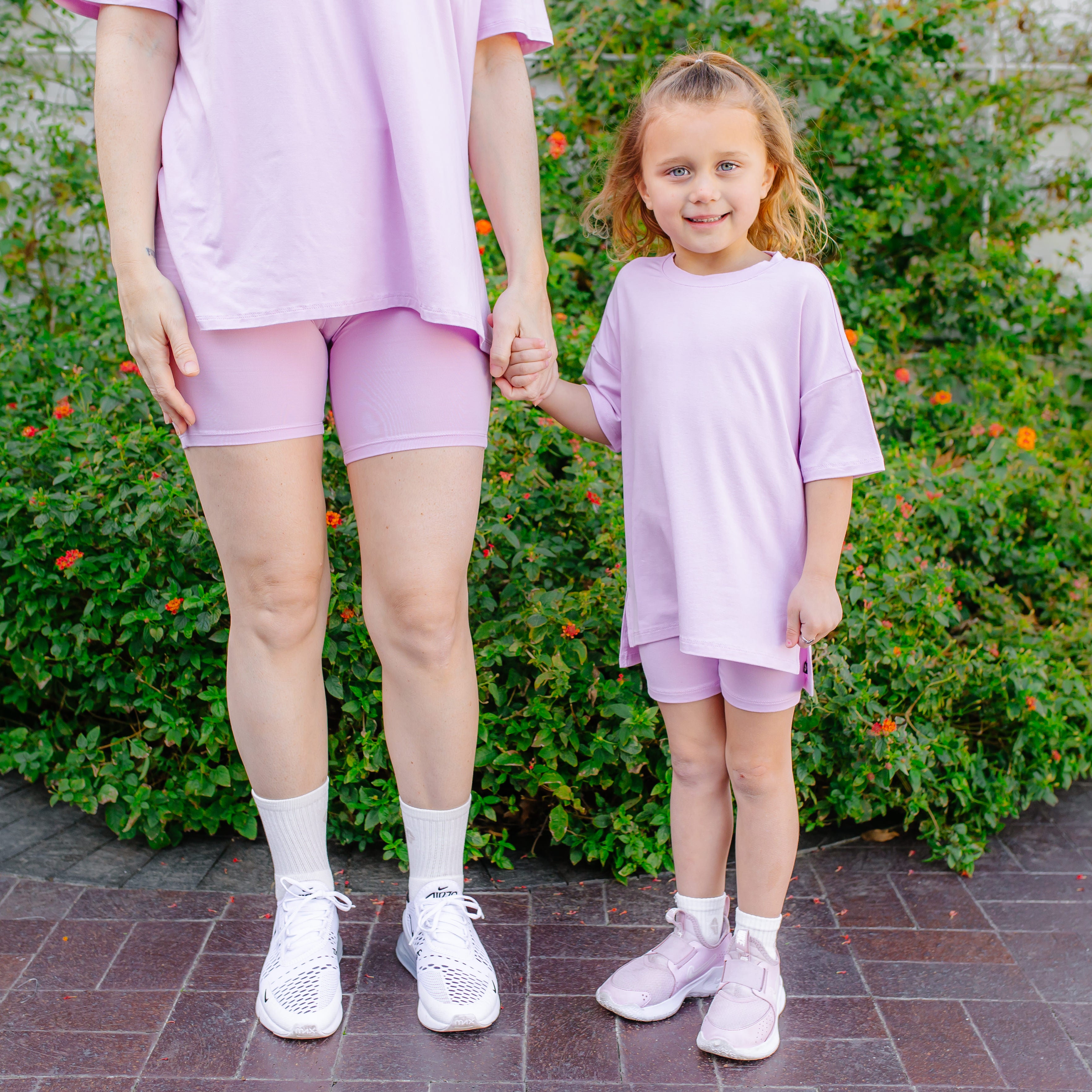 Mom and child in matching purple outfits standing outdoors with greenery in the background