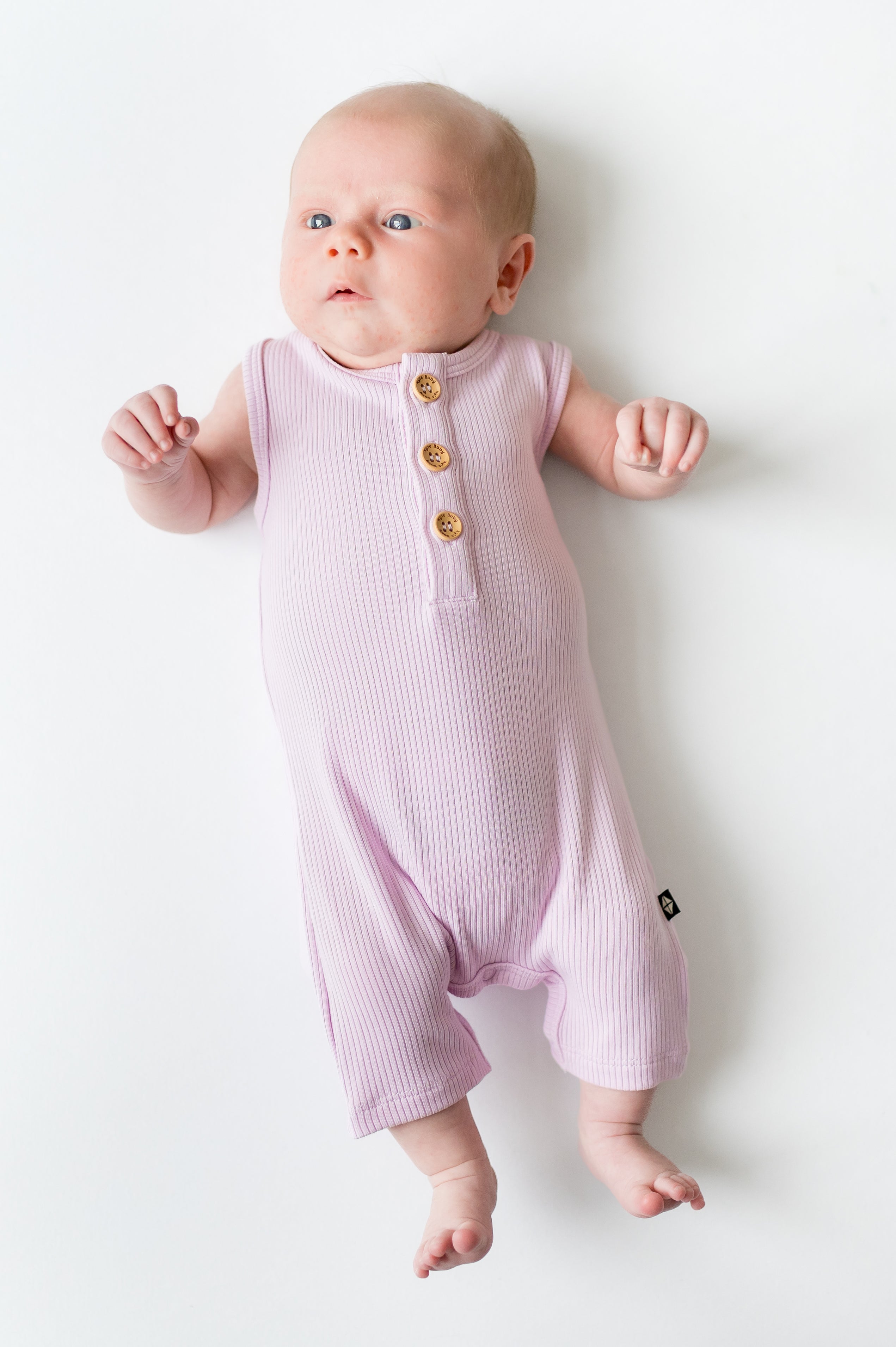 Newborn laying on a white surface wearing the Ribbed Sleeveless Shortall in Thistle