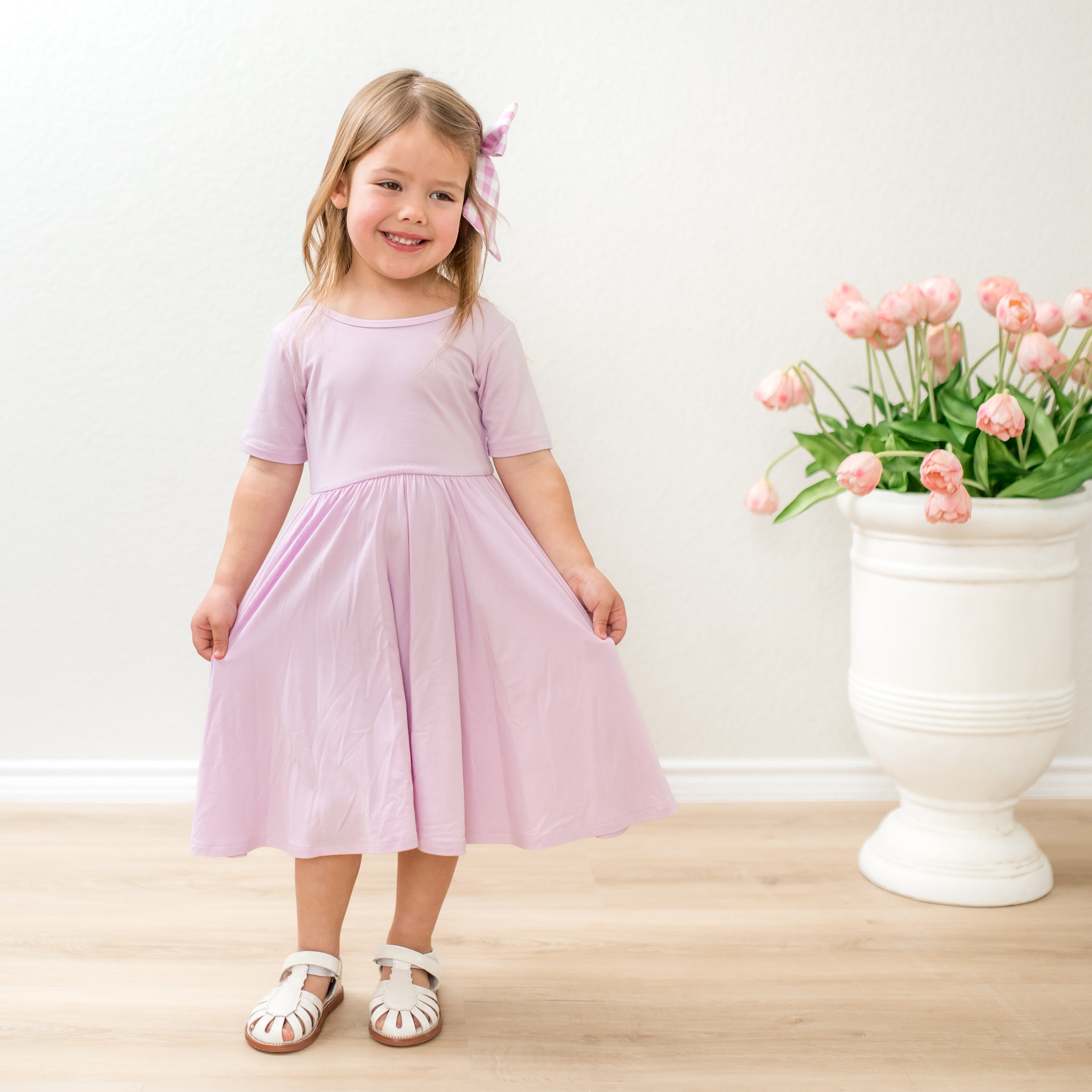 Young girl in a light purple dress standing next to a white vase with pink flowers on a wooden floor.