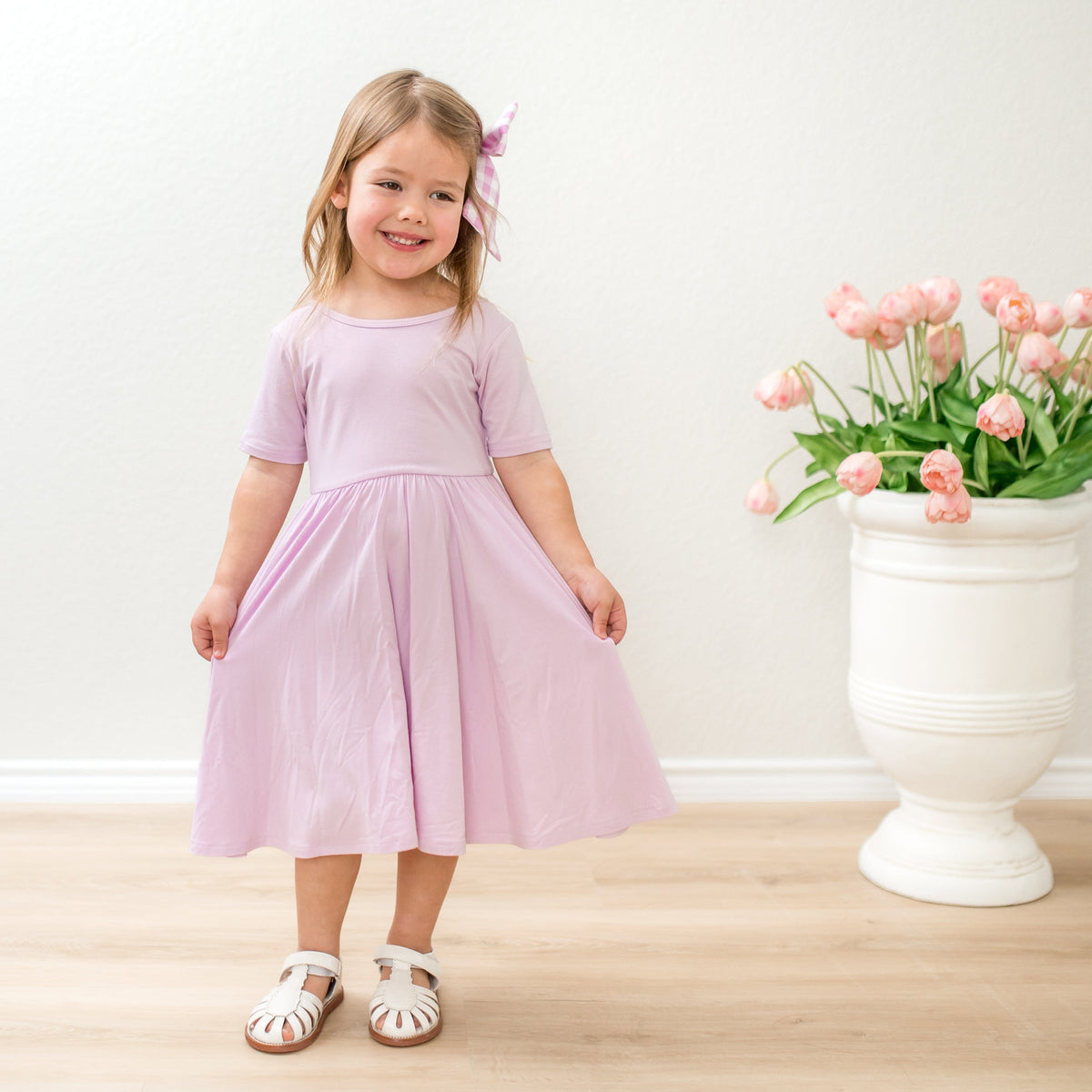 Young girl in a light purple dress standing next to a white vase with pink flowers on a wooden floor.