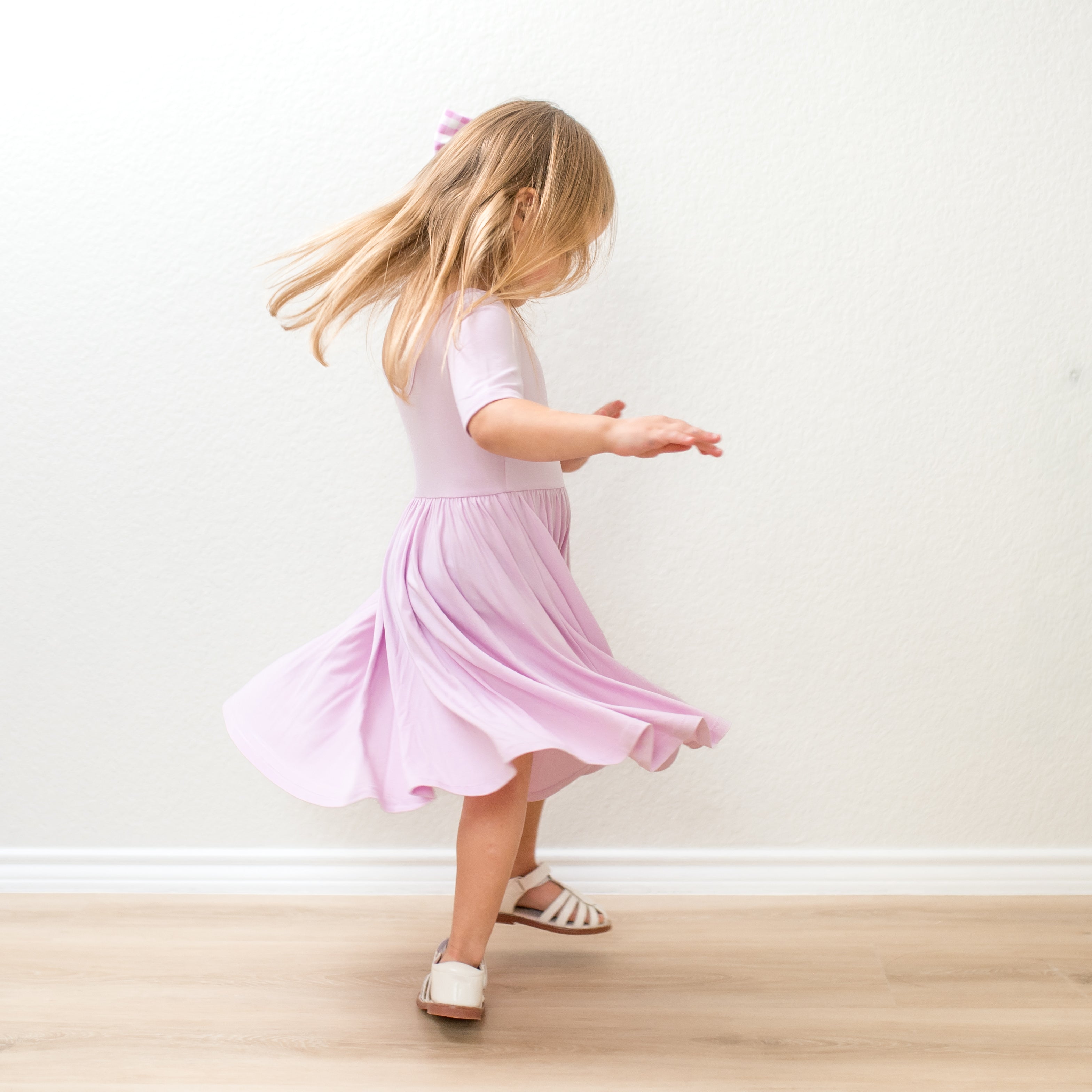 Young girl in a purple dress twirling on a wooden floor against a white wall.