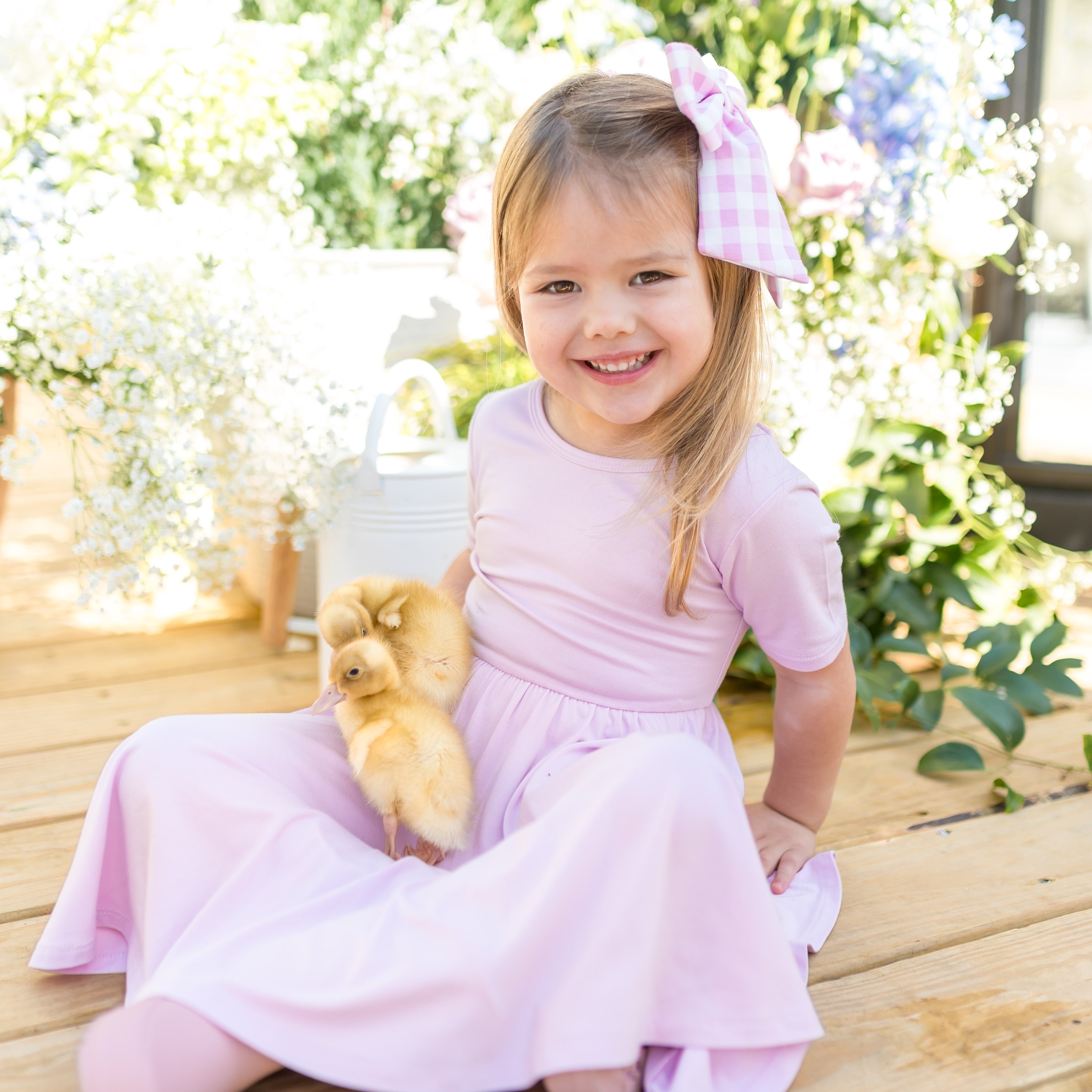 Young girl in a purple dress holding chicks, sitting on a wooden bench with flowers in the background.