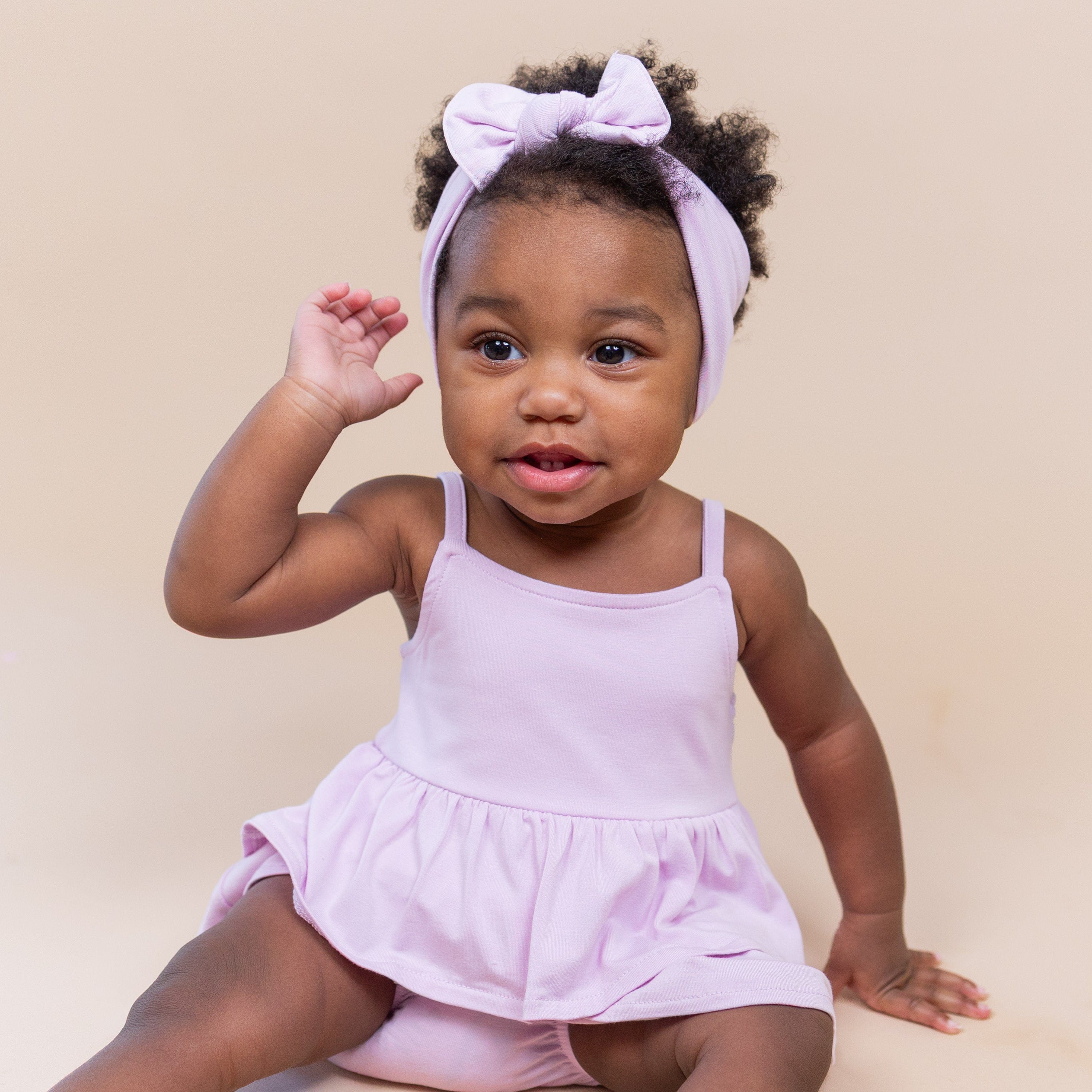 Young toddler sitting on the floor wearing the Spaghetti Strap Ruffle Romper in Thistle with matching bow