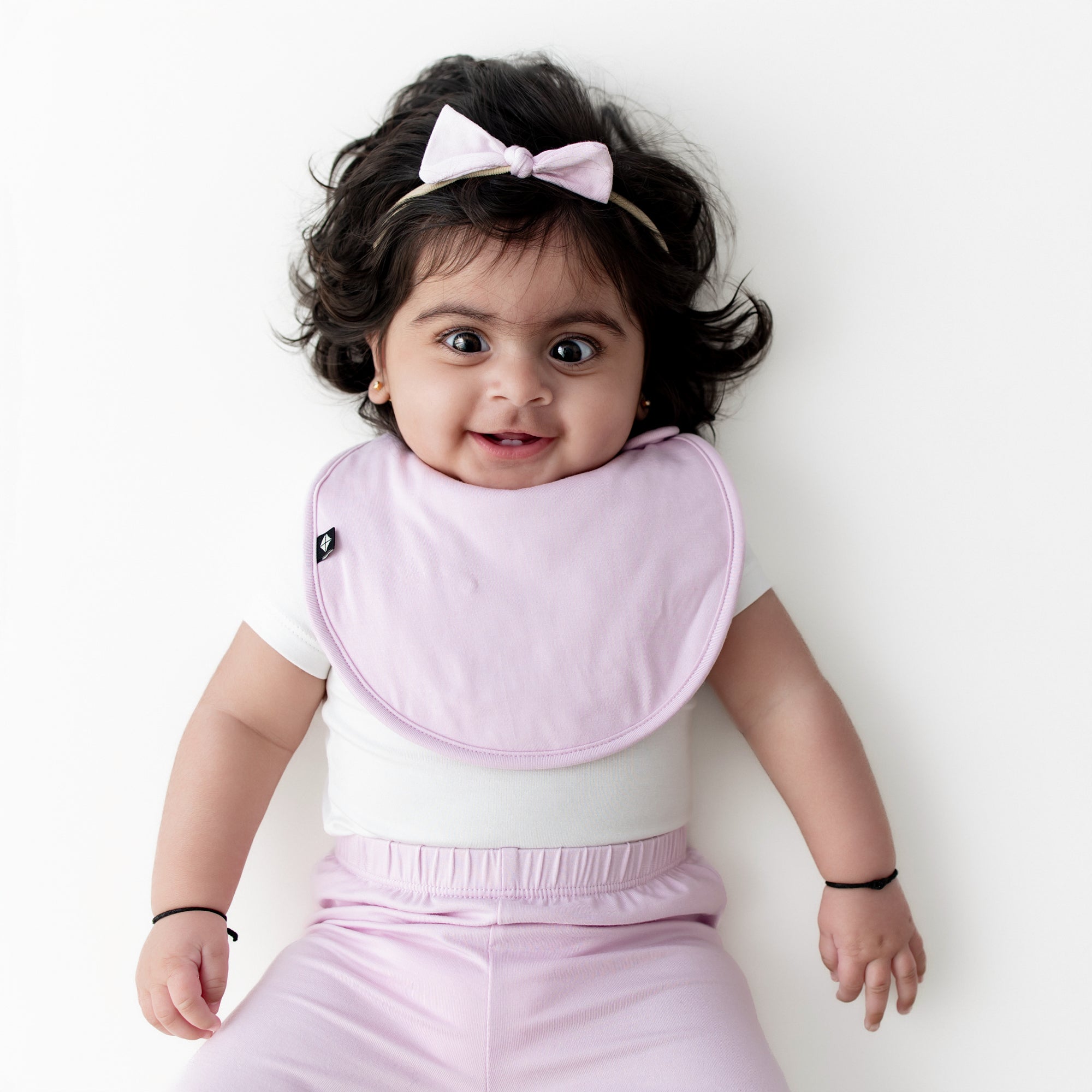 Infant laying on a white surface wearing the Tiny Nylon Bow in Thistle with matching cushy bib and pants
