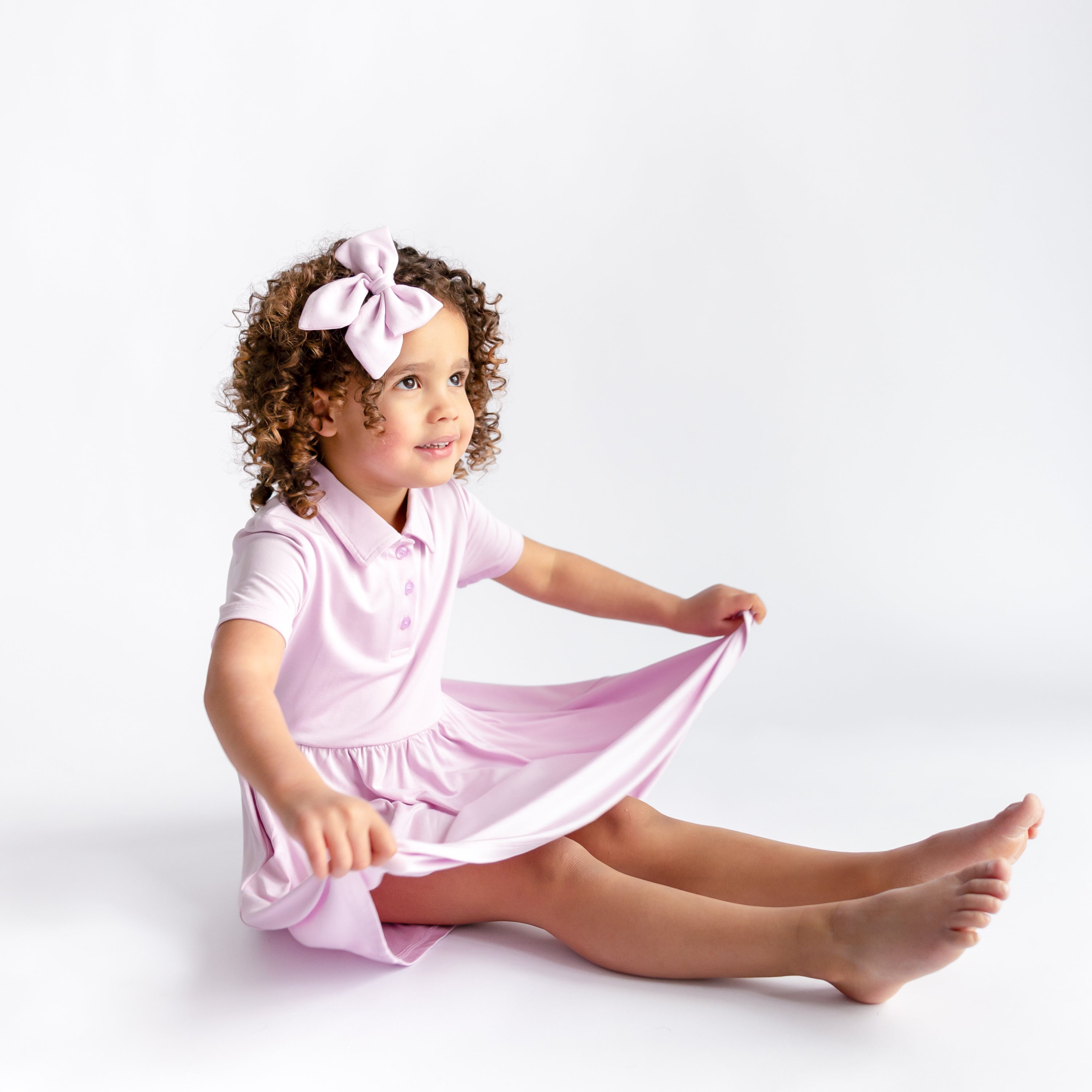 Young girl sitting on the floor wearing the Polo Dress in Thistle with matching bow