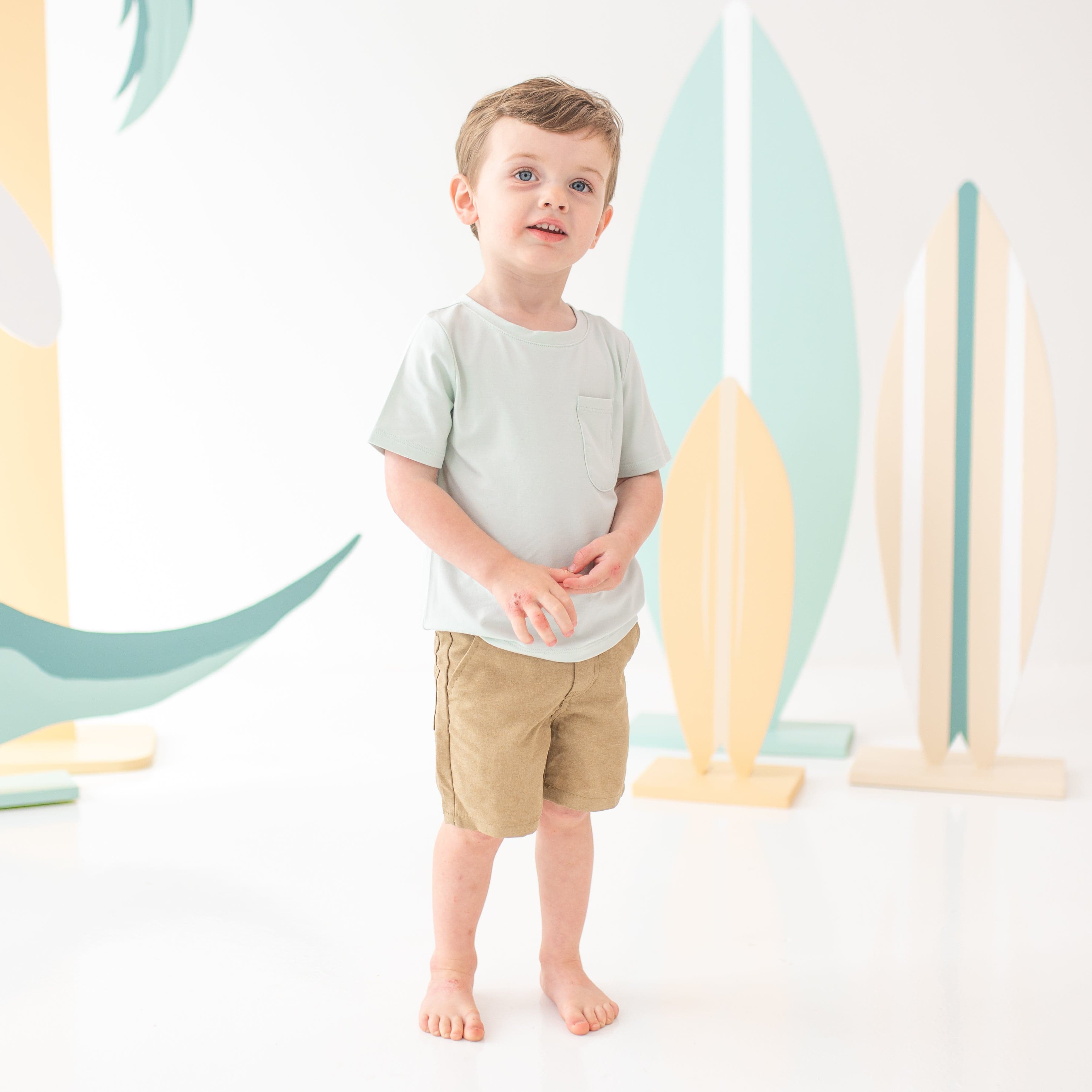 Young child model standing holding his hands together wearing the Toddler Crew Neck Tee in Dew with tan shorts