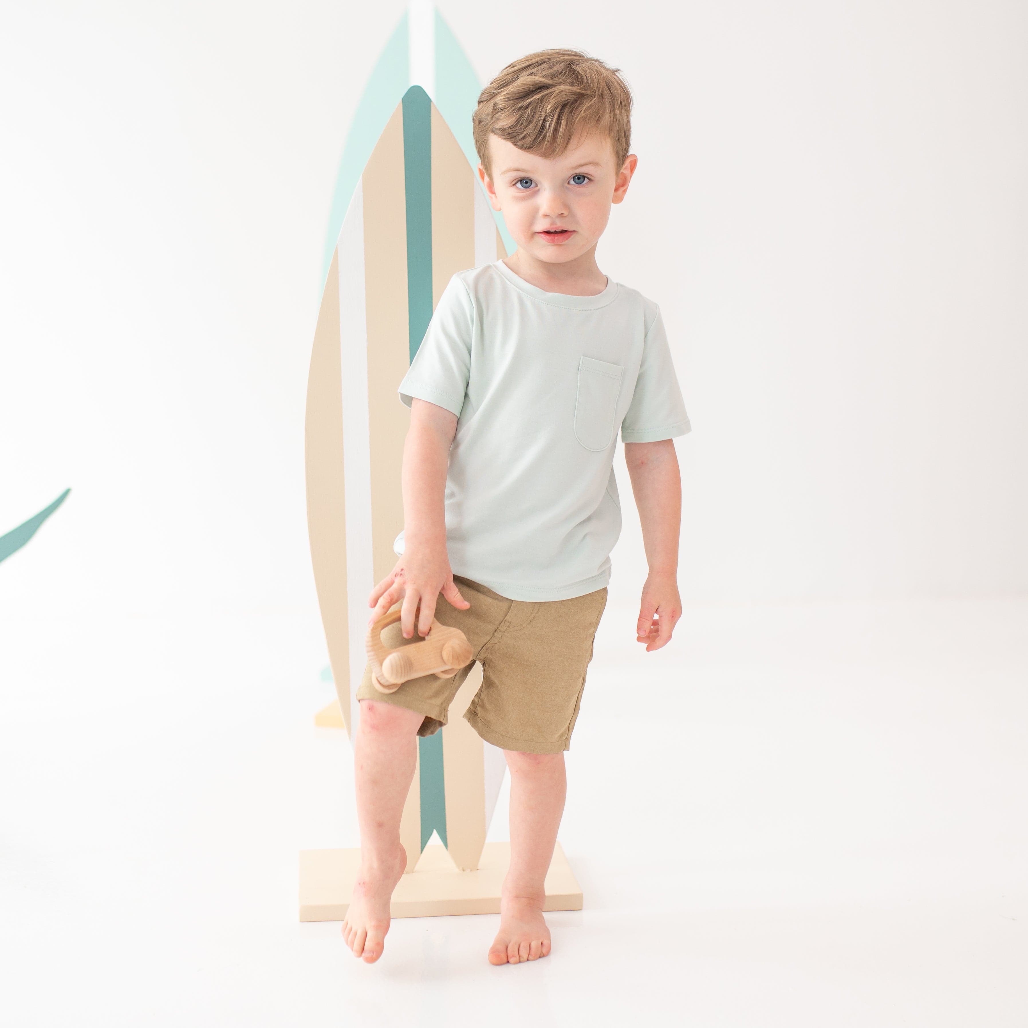 Young boy model standing on one foot holding a wooden car wearing the Toddler Crew Neck Tee in Dew with tan shorts