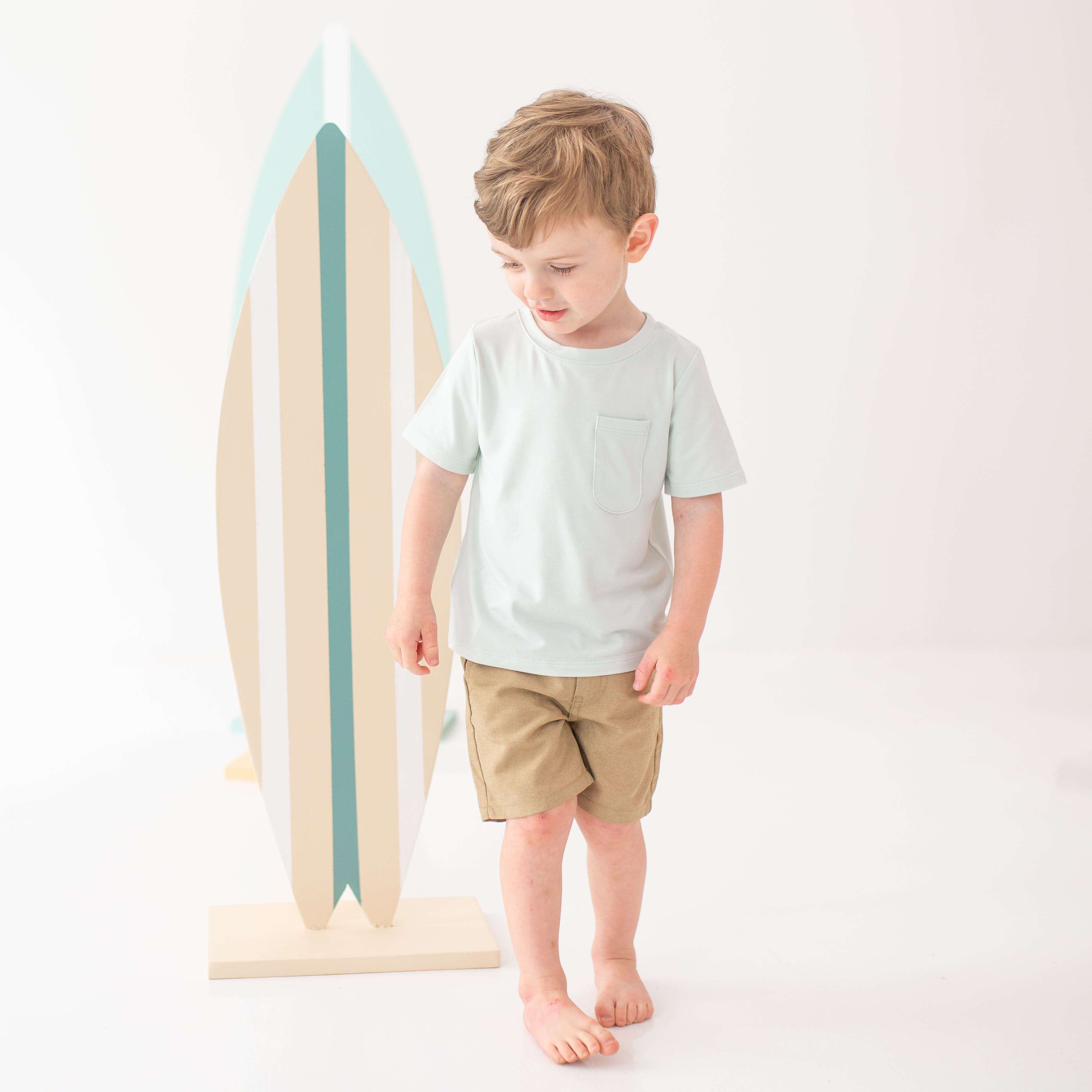 Young boy model looking down and walking wearing the Toddler Crew Neck Tee in Dew with tan shorts