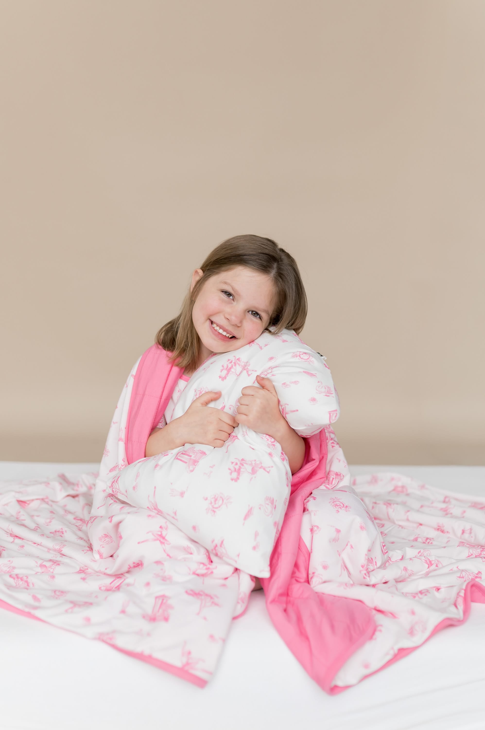 Young smiling girl sitting with the Youth Blanket in Toile Ballet 1.0 wrapped around her shoulders holding a pillow with a toile ballet pillow case on it