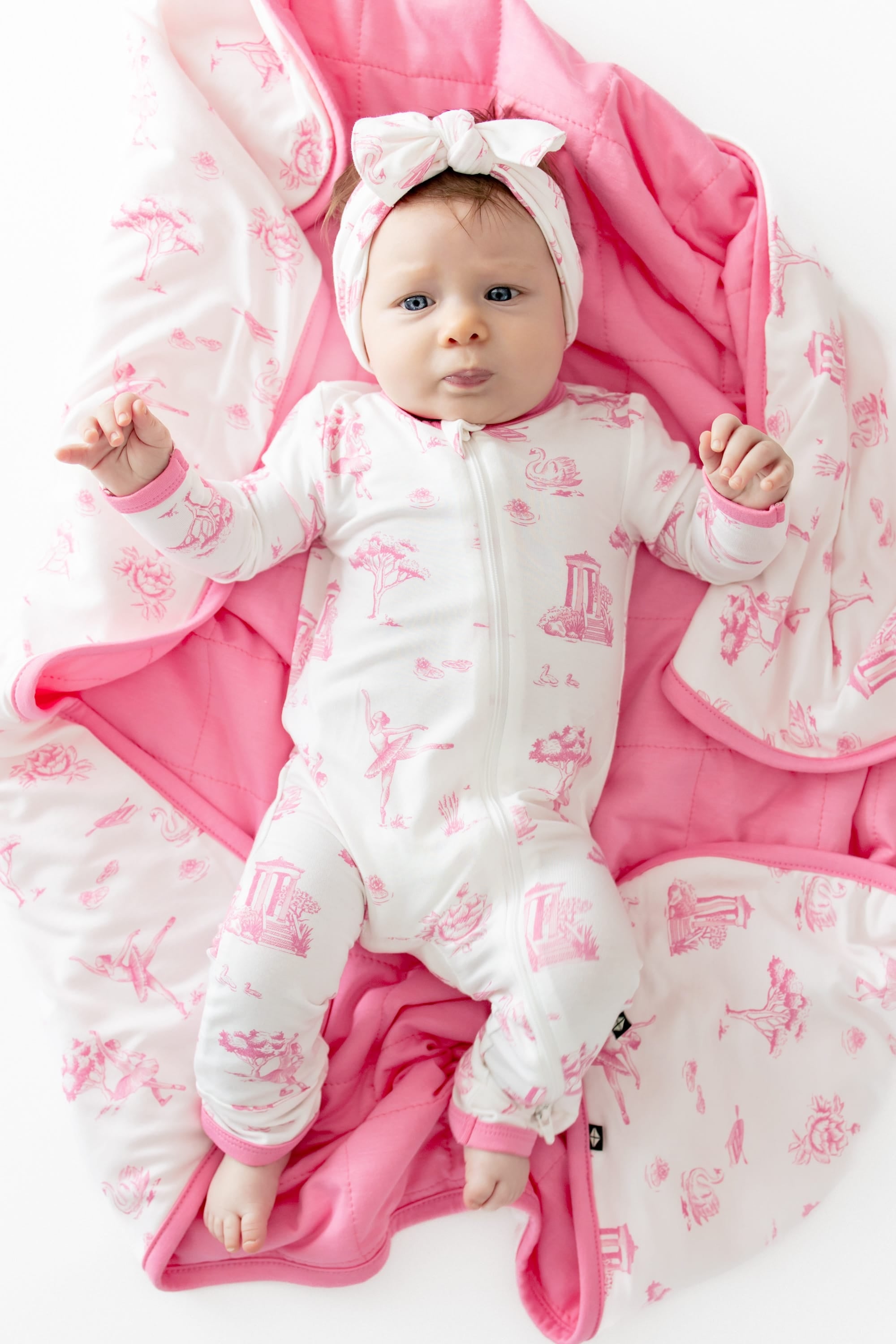 Close up of infant laying on a Baby Blanket in Toile Ballet wearing a matching zippered romper and knotted bow