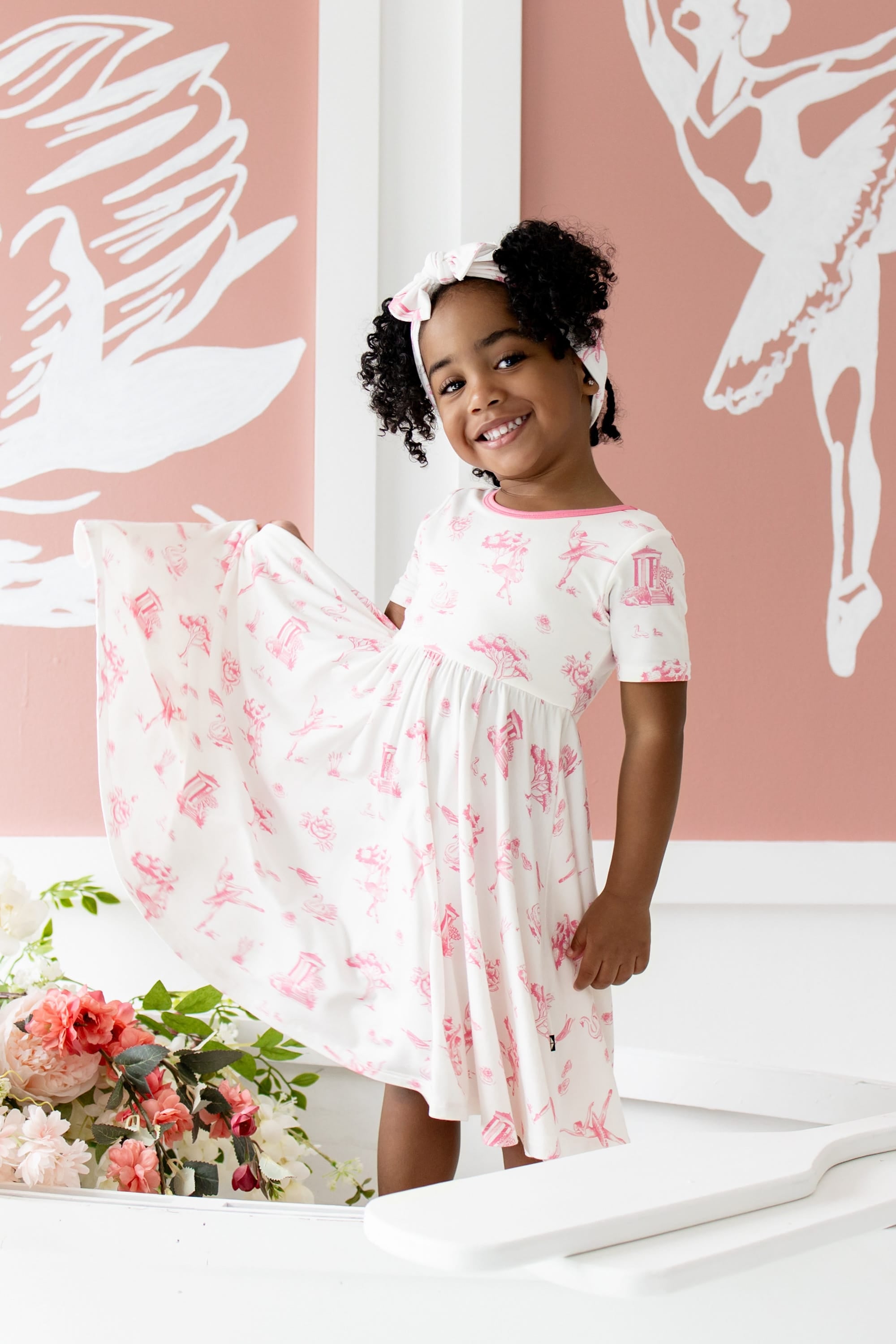 Young girl posing wearing the Twirl Dress in Toile Ballet standing in a white wooden rowboat beside flowers in front of ballet themed art