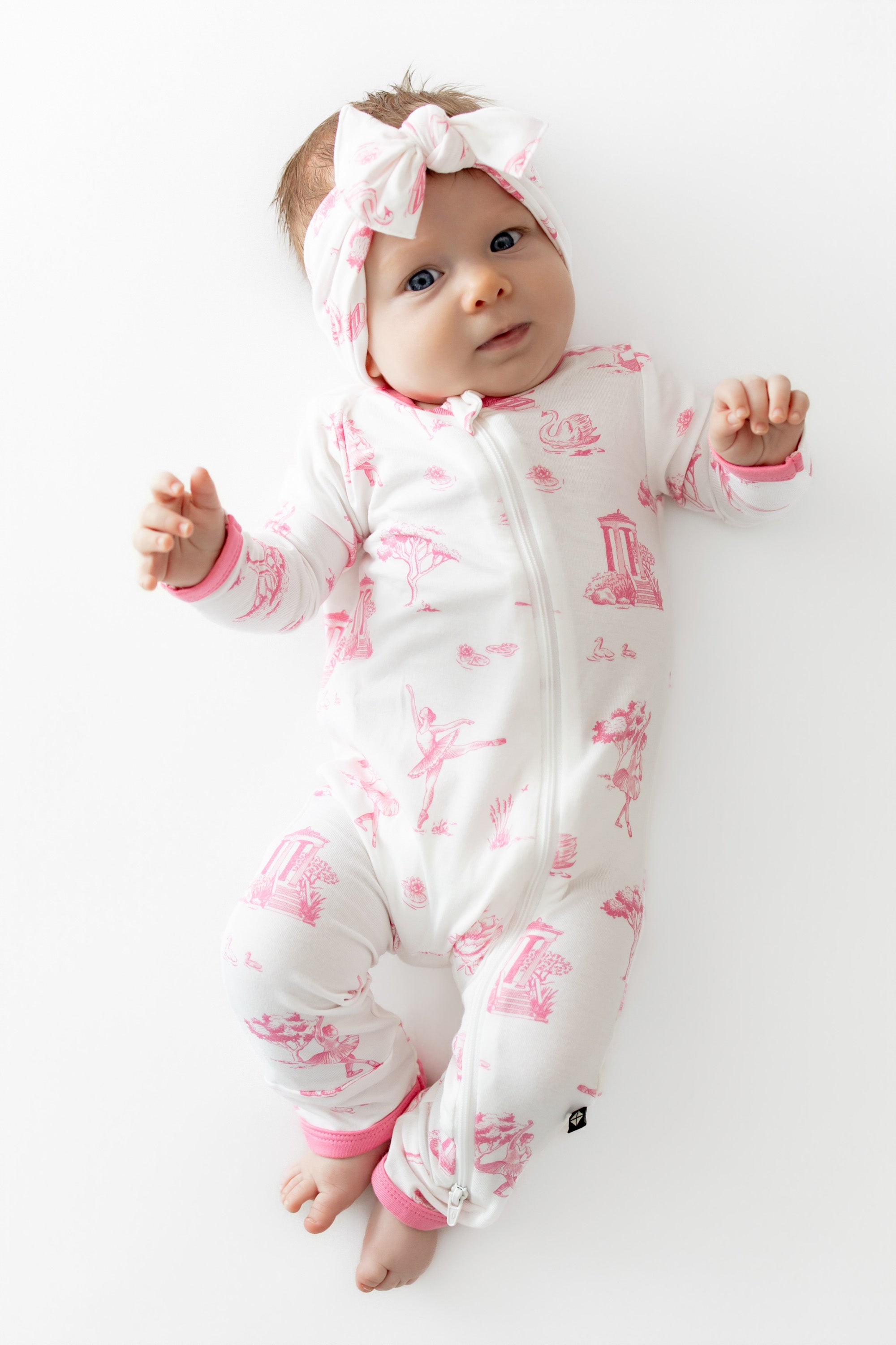 Infant laying on a white surface wearing the Zippered Romper in Toile Ballet and matching bow