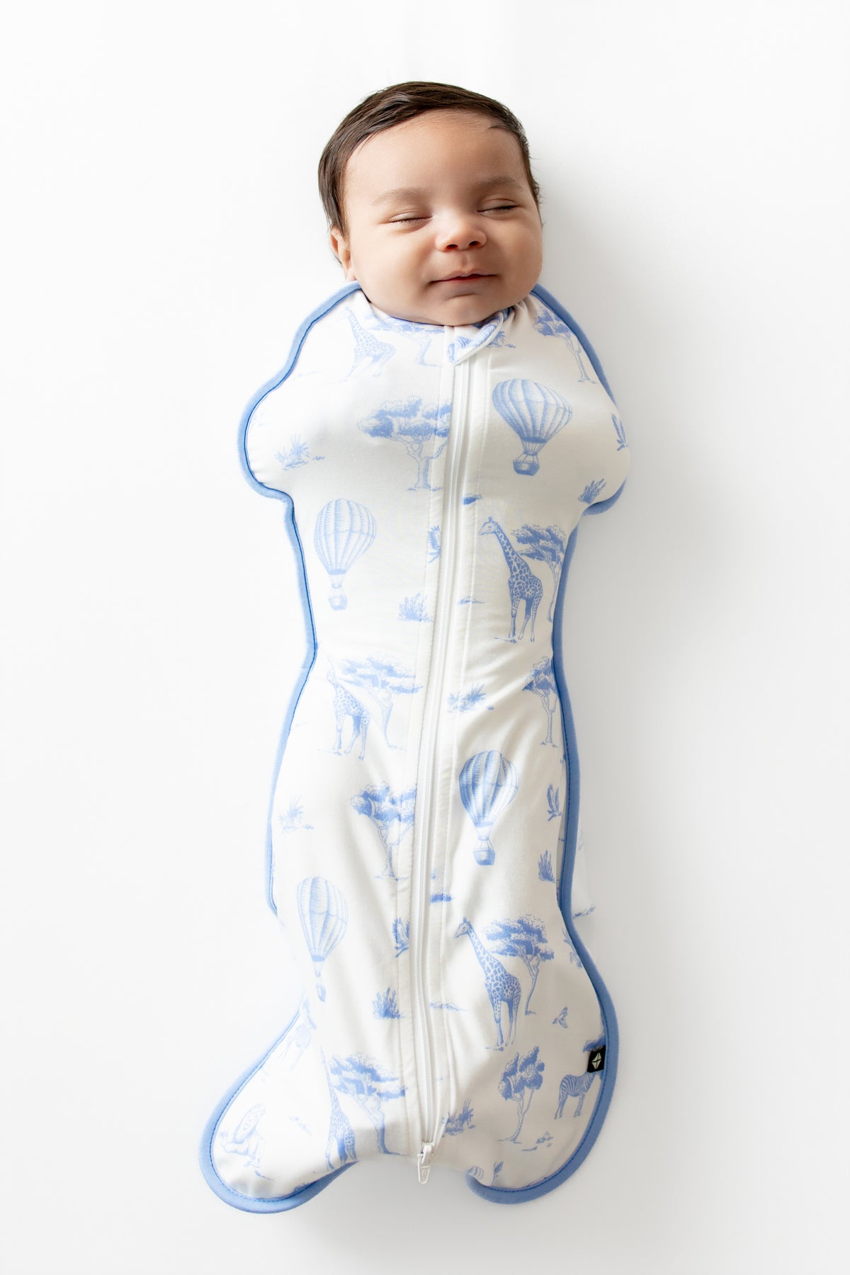 Newborn laying on a white surface wearing the Swaddle Bag in Toile Safari 0.5