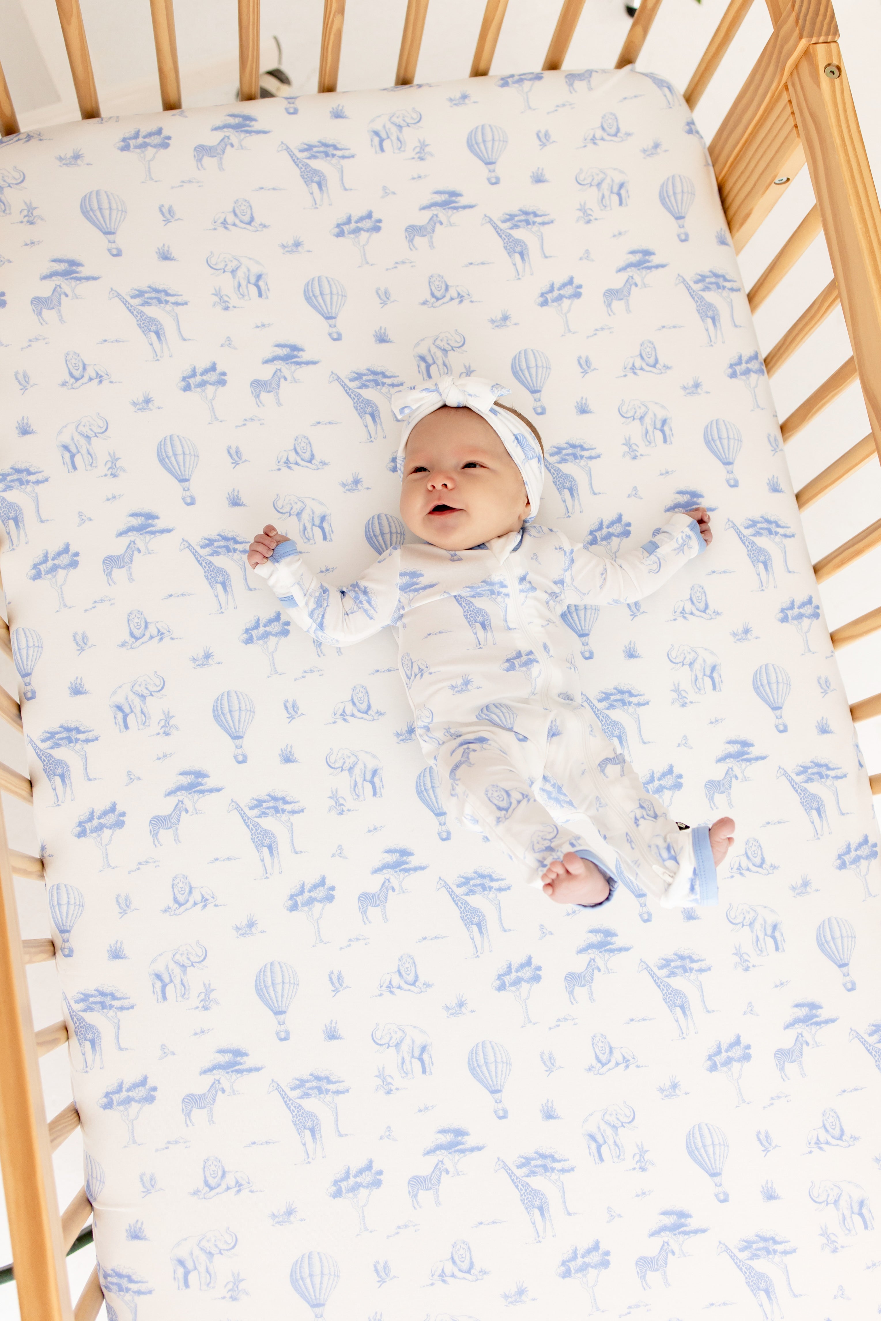 Infant laying in a crib on a Crib Sheet in Toile Safari wearing the matching zippered romper and bow
