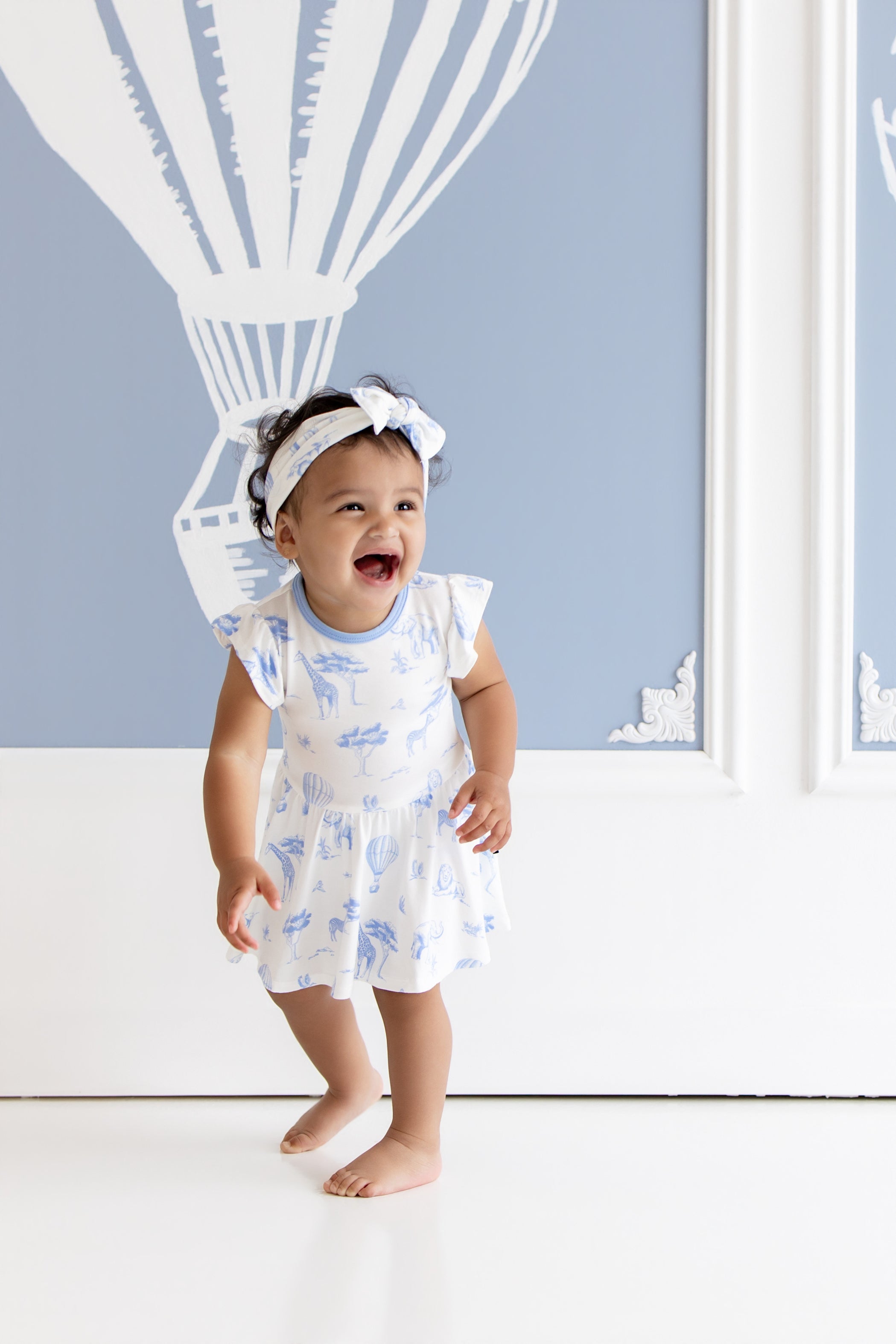 Young toddler laughing wearing the Twirl Bodysuit Dress in Toile Safari standing in front of blue and white safari pictures