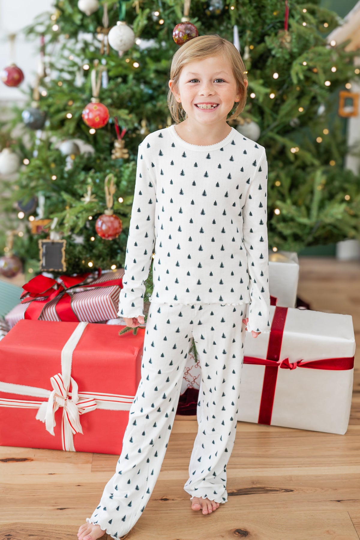 A young girl stands barefoot in front of a decorated Christmas tree and wrapped gift boxes, wearing a white long-sleeve pointelle set with a small dark pine-tree pattern.