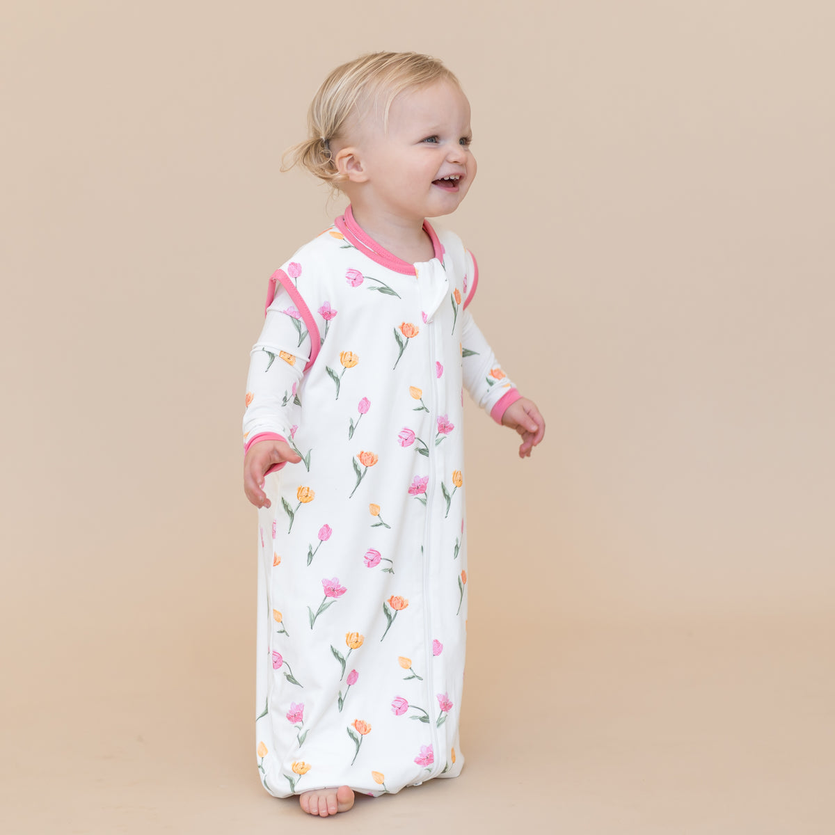 Young smiling toddler standing wearing the Sleep Bag Walker in Tulip 0.5
