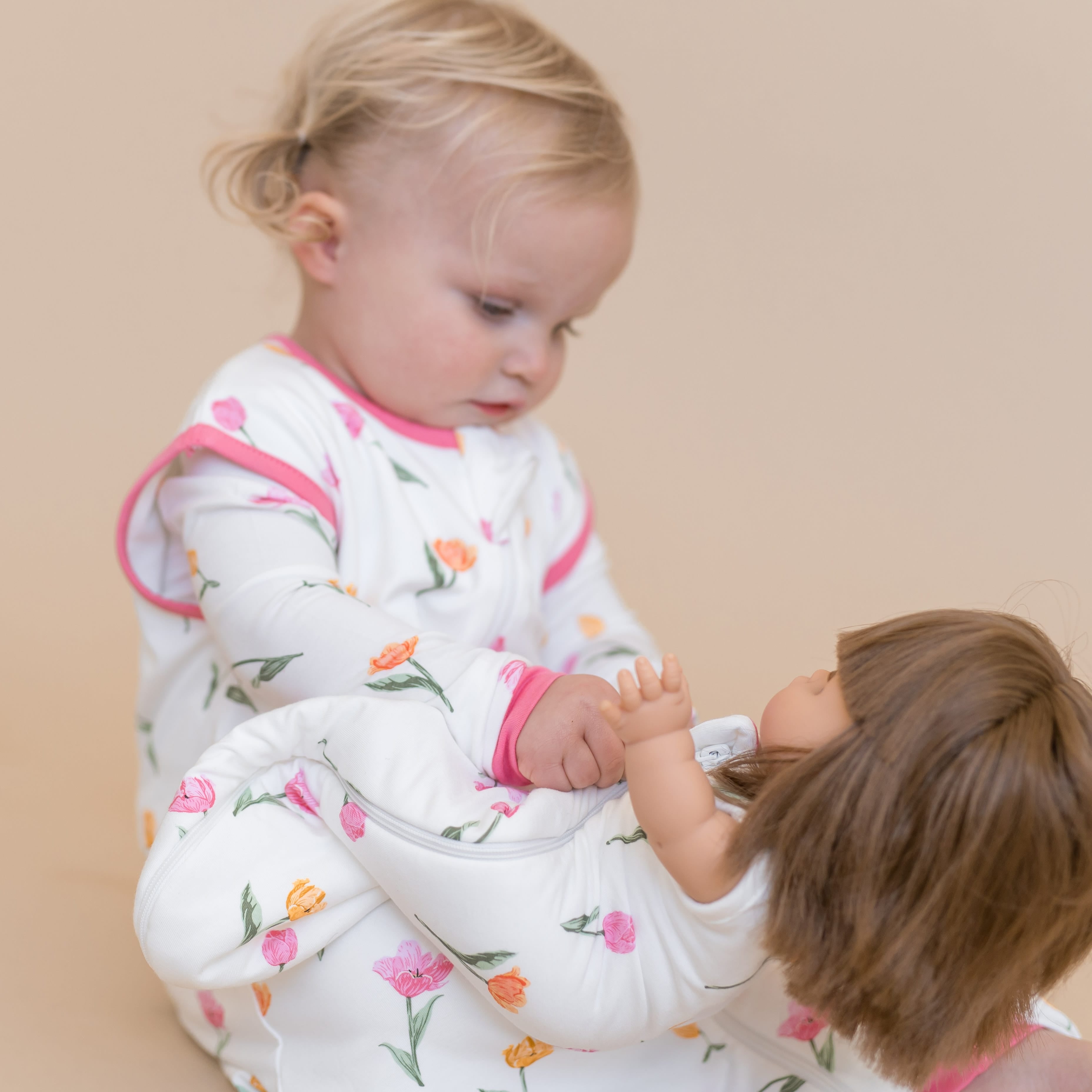 Young toddler sitting on the floor wearing the Sleep Bag Walker in Tulip 0.5 playing with a doll wearing a matching doll sleep bag