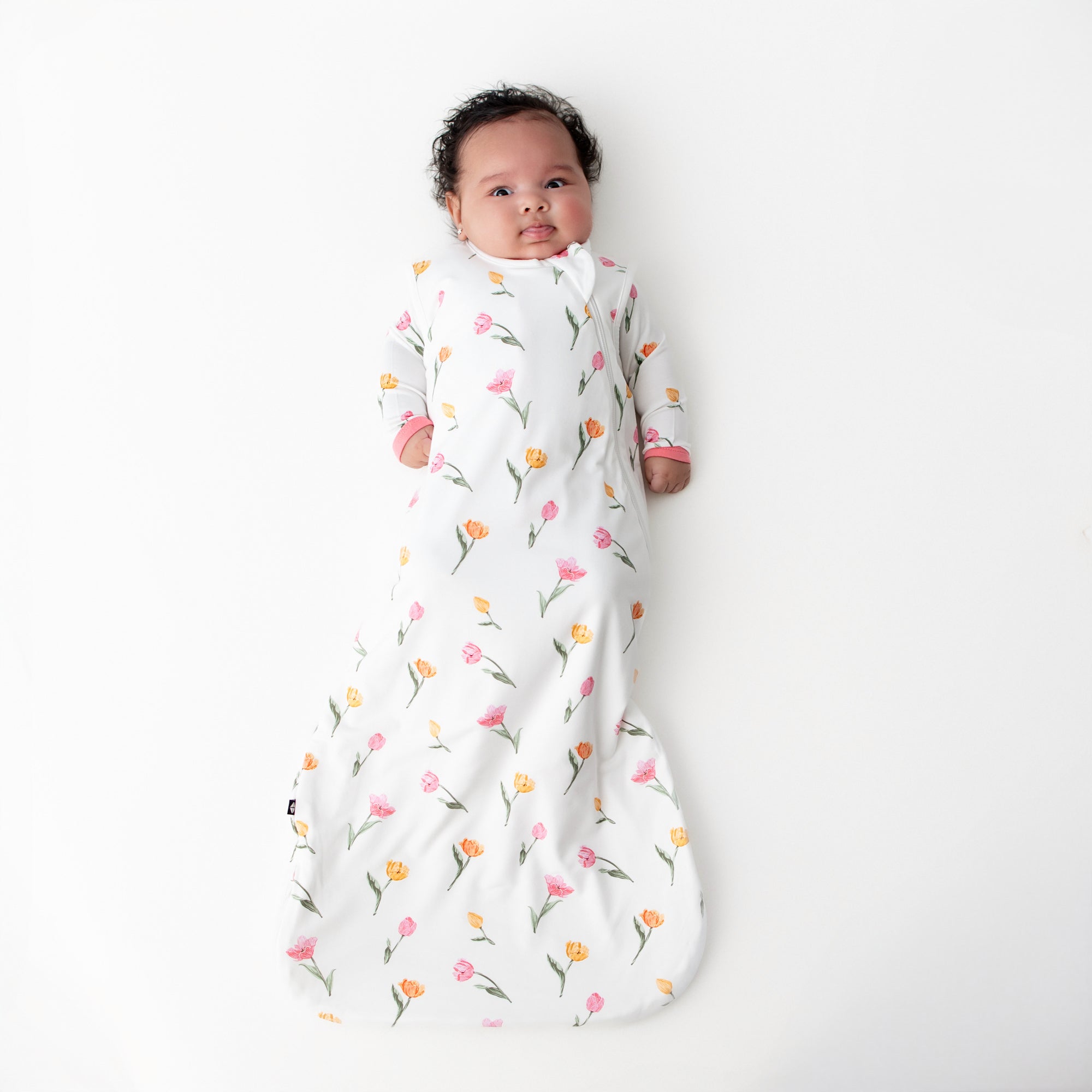 Young infant laying on a white surface wearing the Sleep Bag in Tulip 1.0 with a matching romper underneath