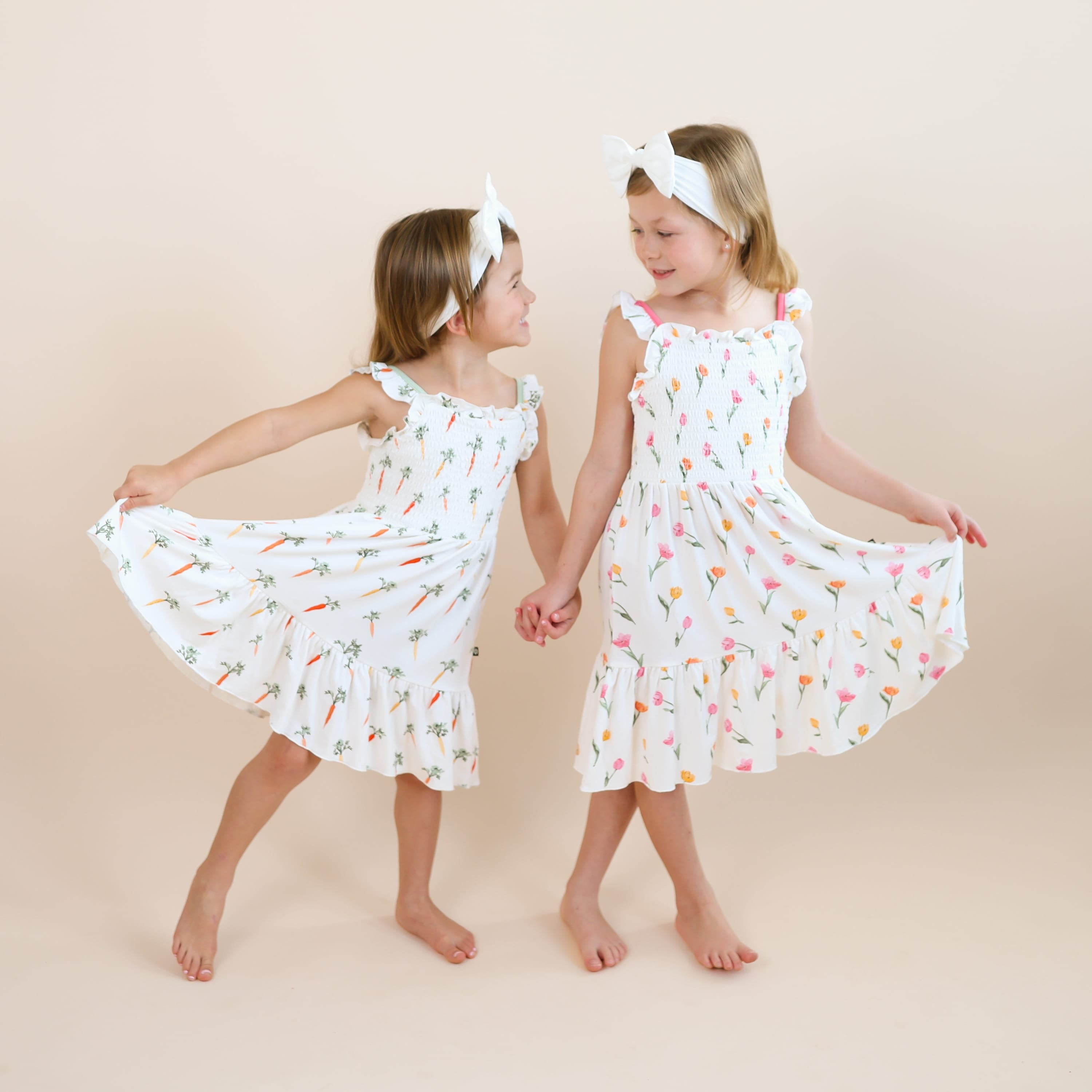 Two sisters holding hands looking at one another wearing the Smocked Dress in Carrot and Tulip