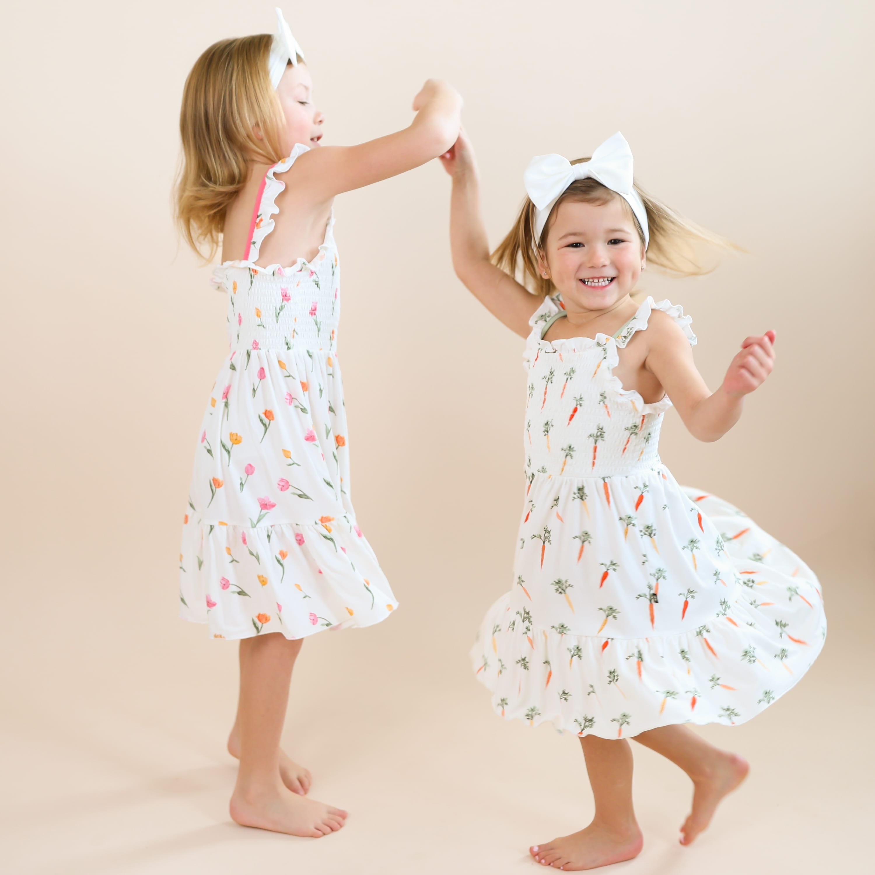 Two sisters holding hands with one of them twirling in a circle both wearing the Smocked Dress in Tulip and Carrot