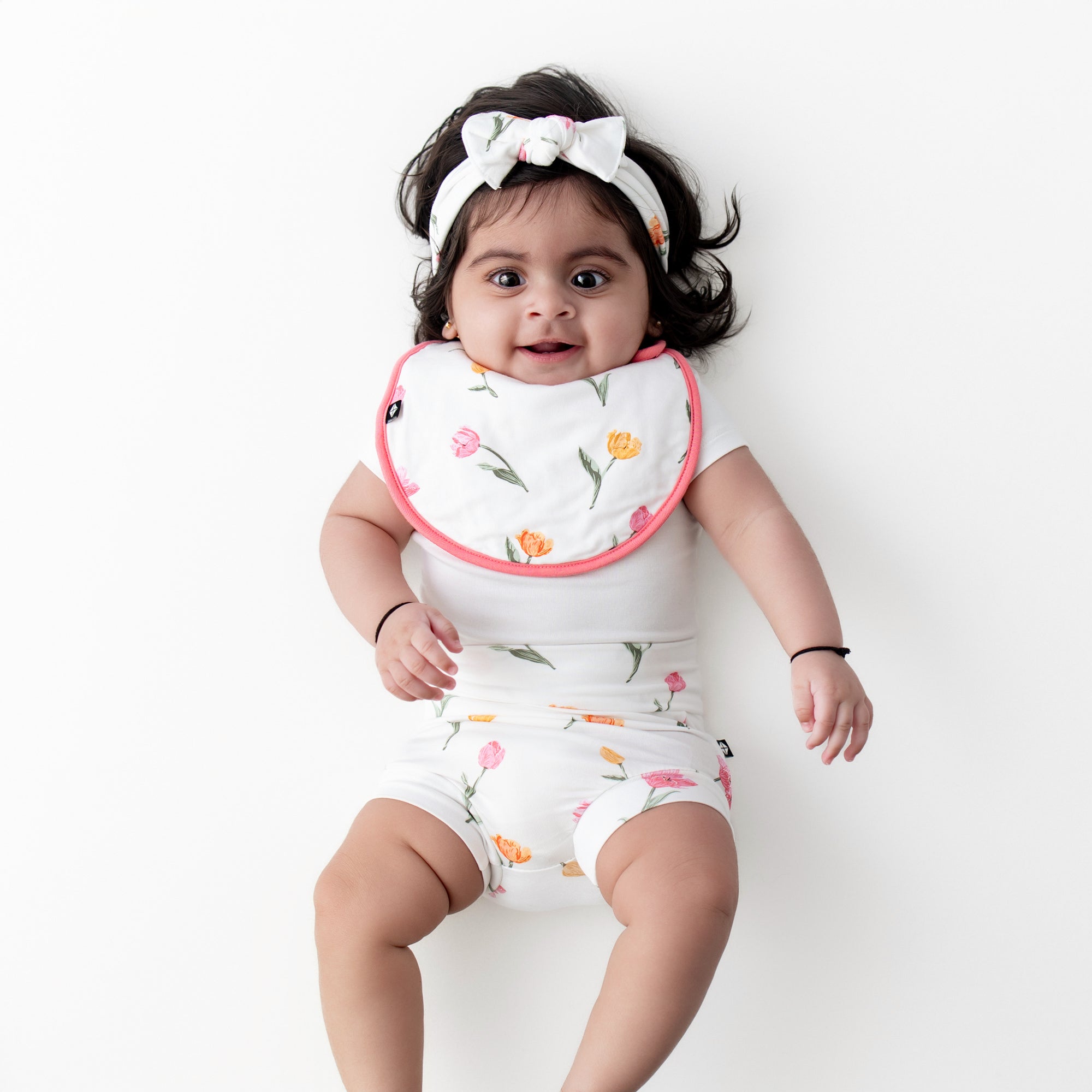 Infant wearing the Bummies, Cushy Bib and knotted bow headband in Tulip paired with a white short sleeve bodysuit