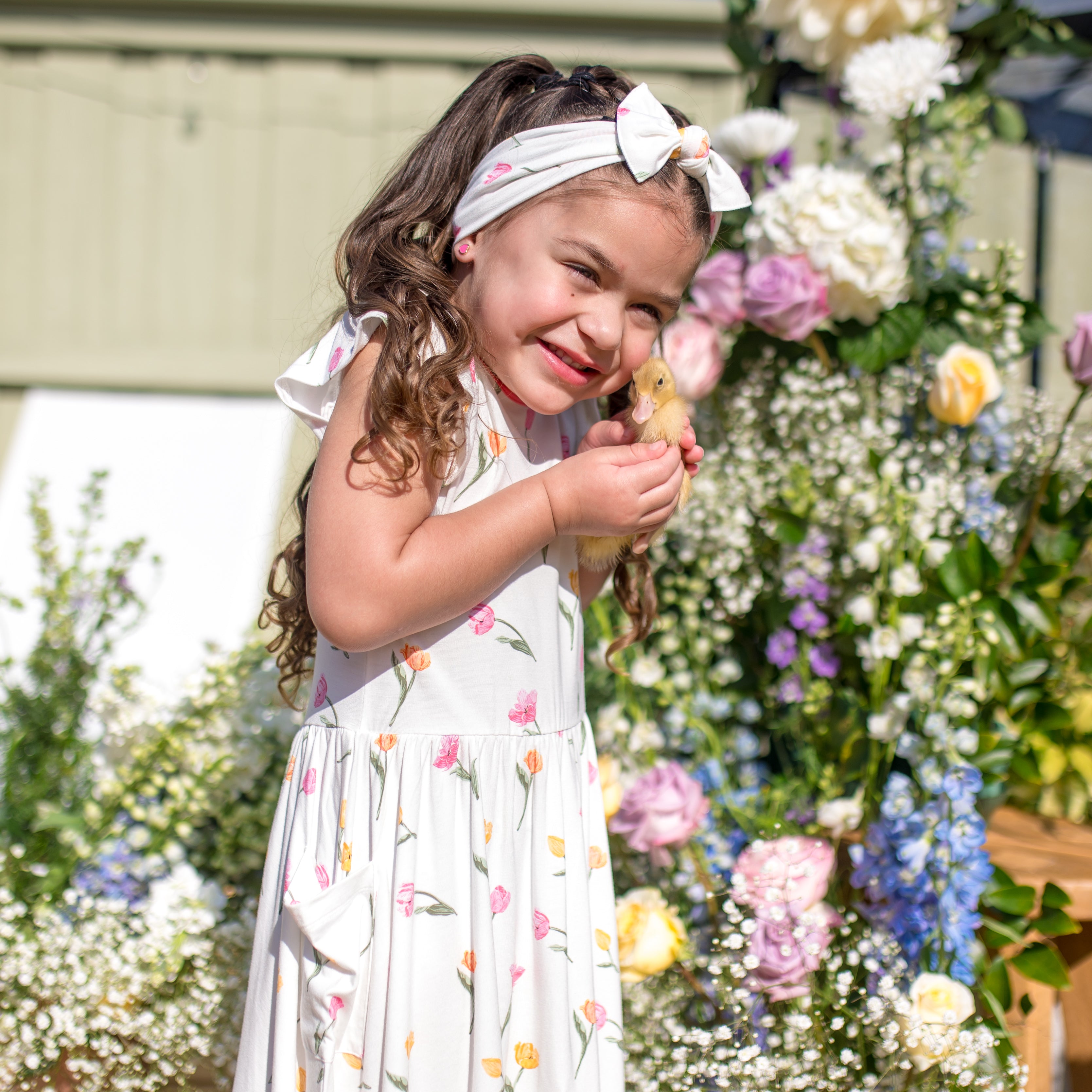 Young girl standing in front of flowers wearing the Pocket Dress in Tulip holding a small duck