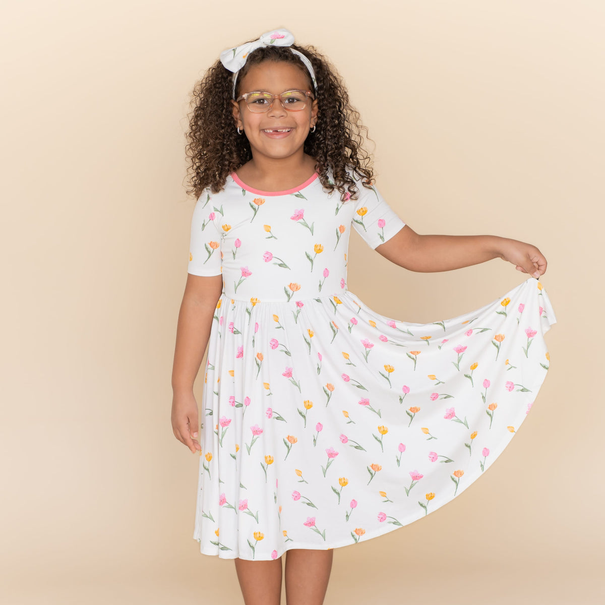 Young girl standing in front of a light taupe wall wearing the Twirl Dress in Tulip holding the hem of the skirt up