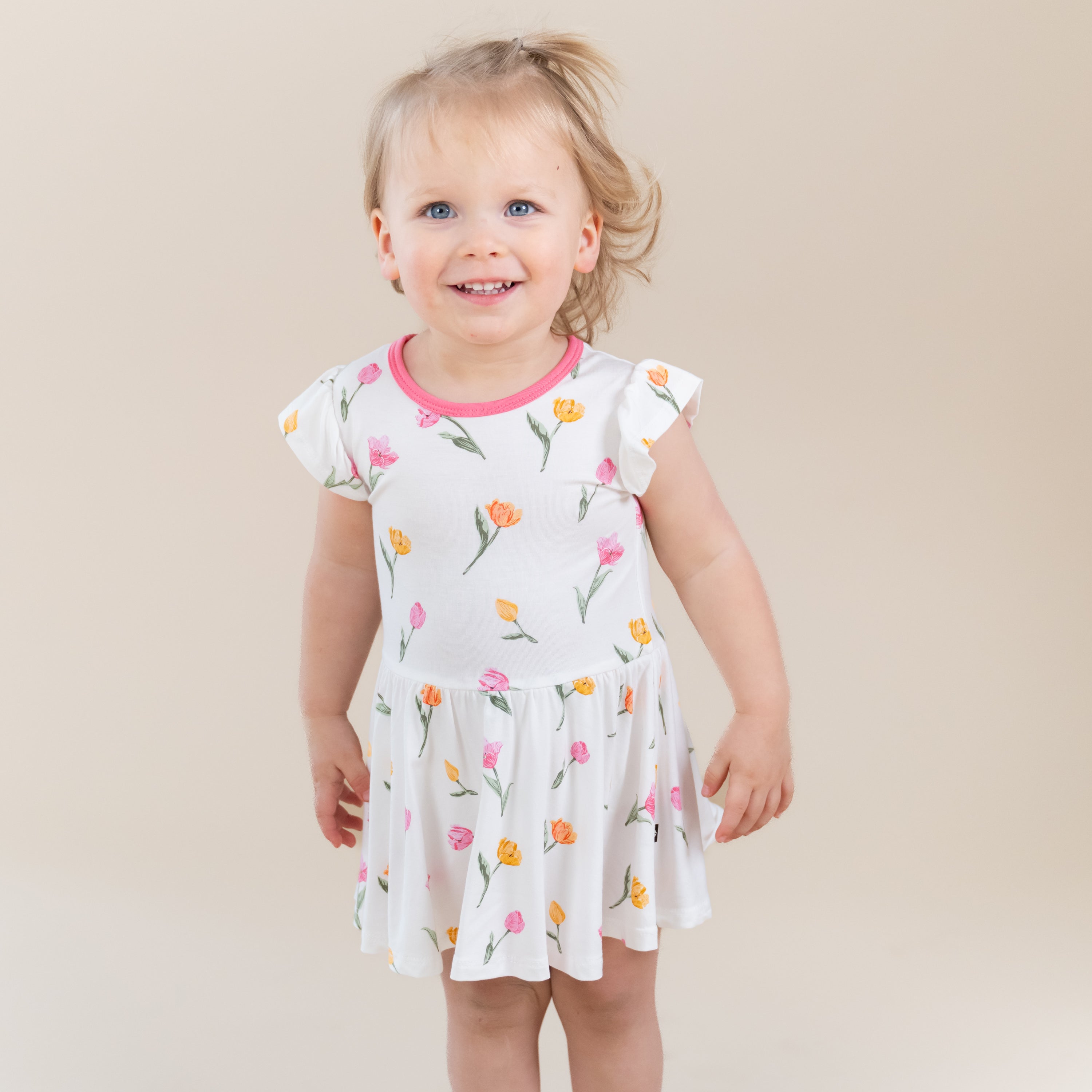 Smiling toddler wearing the Twirl Bodysuit Dress in Tulip standing in front of a light taupe background