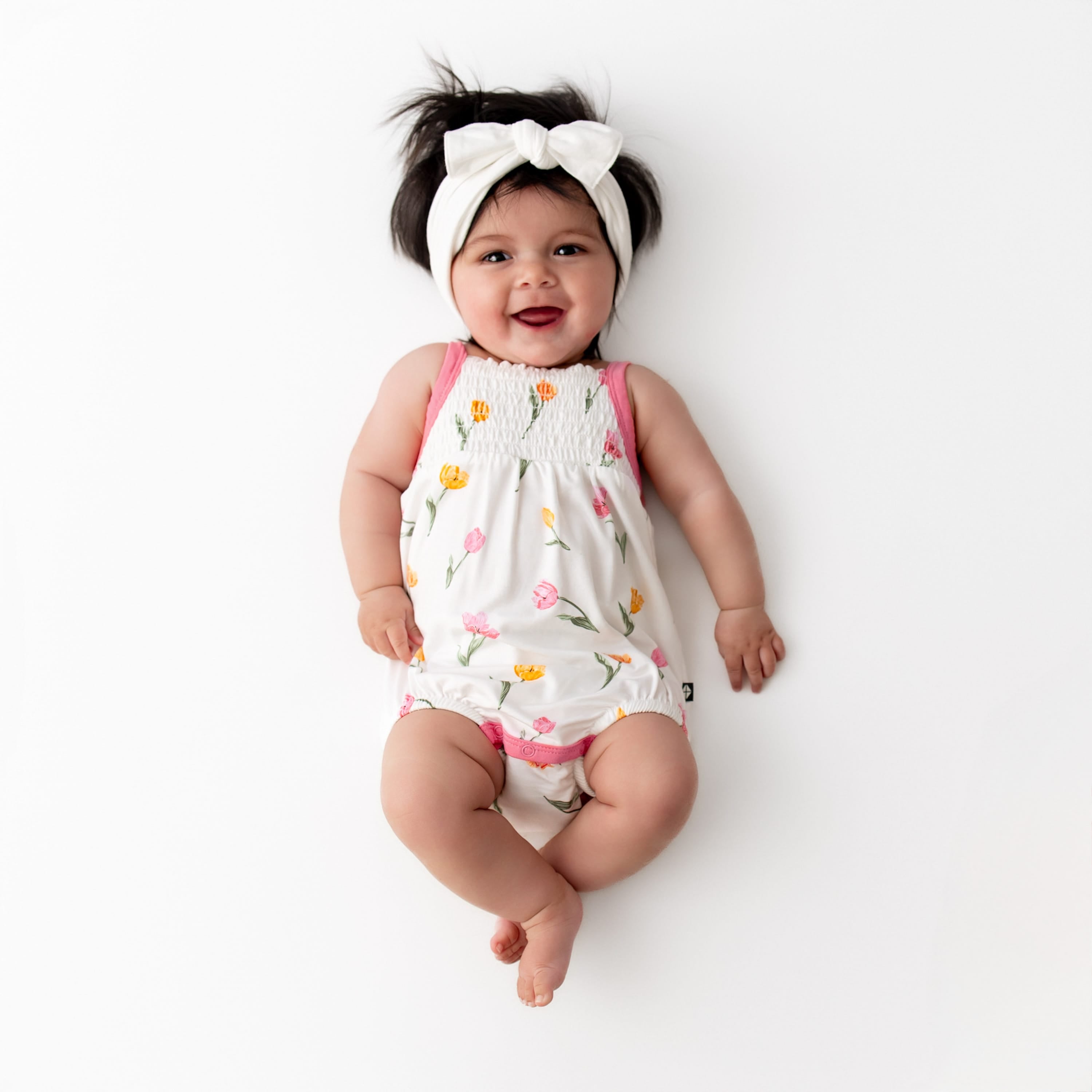 Smiling infant wearing the Smocked Bubble Romper in Tulip with a white knotted bow headband