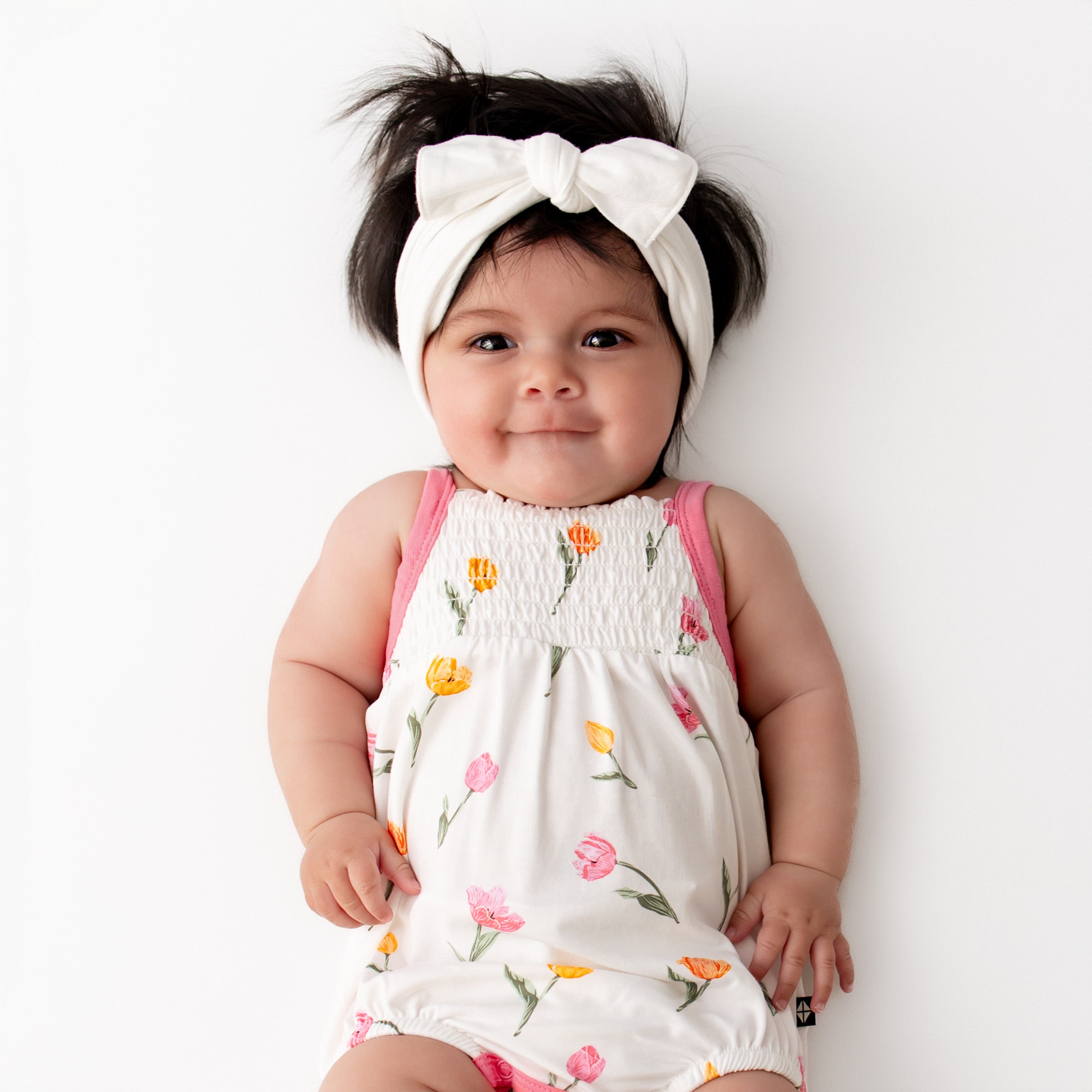 Close up of smiling infant wearing the Smocked Bubble Romper in Tulip with a white knotted bow headband