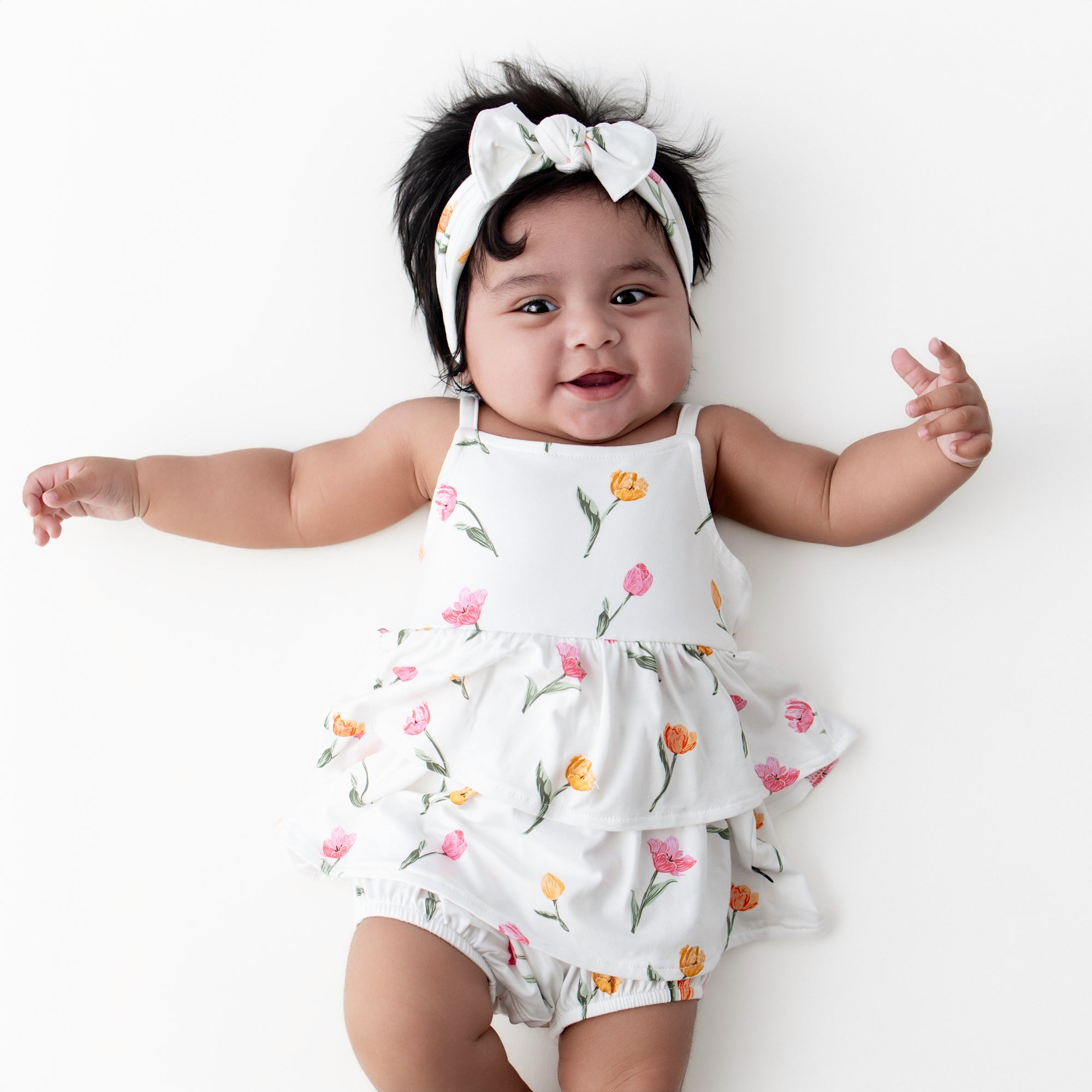 Infant laying on a white surface wearing the Spaghetti Strap Ruffle Romper in Tulip with matching bow headband
