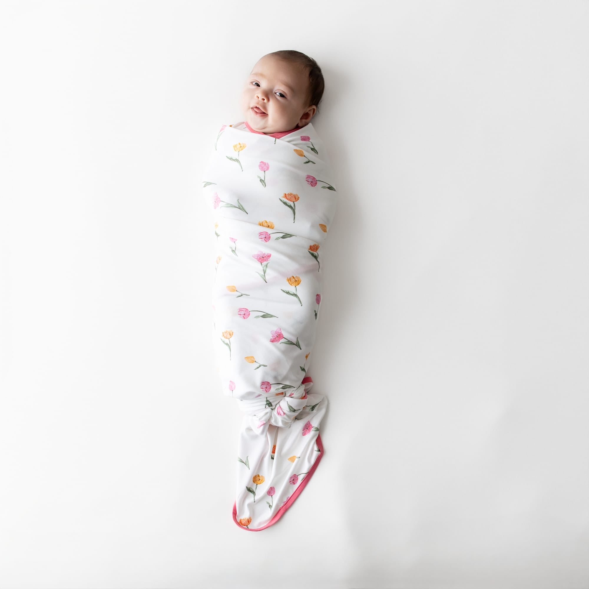 Newborn swaddled in the Swaddle Blanket in Tulip laying on a white surface