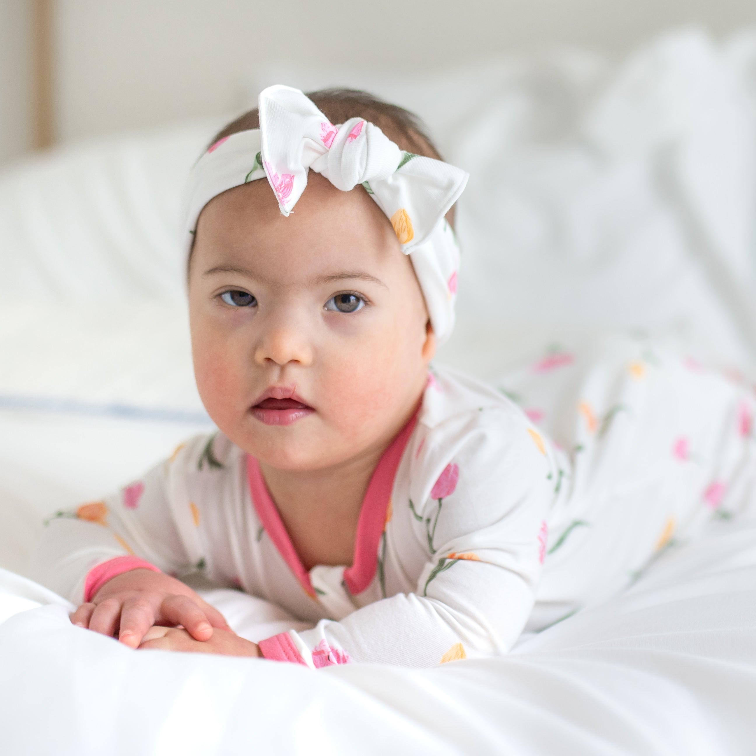 Close up of a toddler laying on a bed on her stomach wearing the Zippered Footie in Tulip showing the pink trim neckline and matching tulip bow