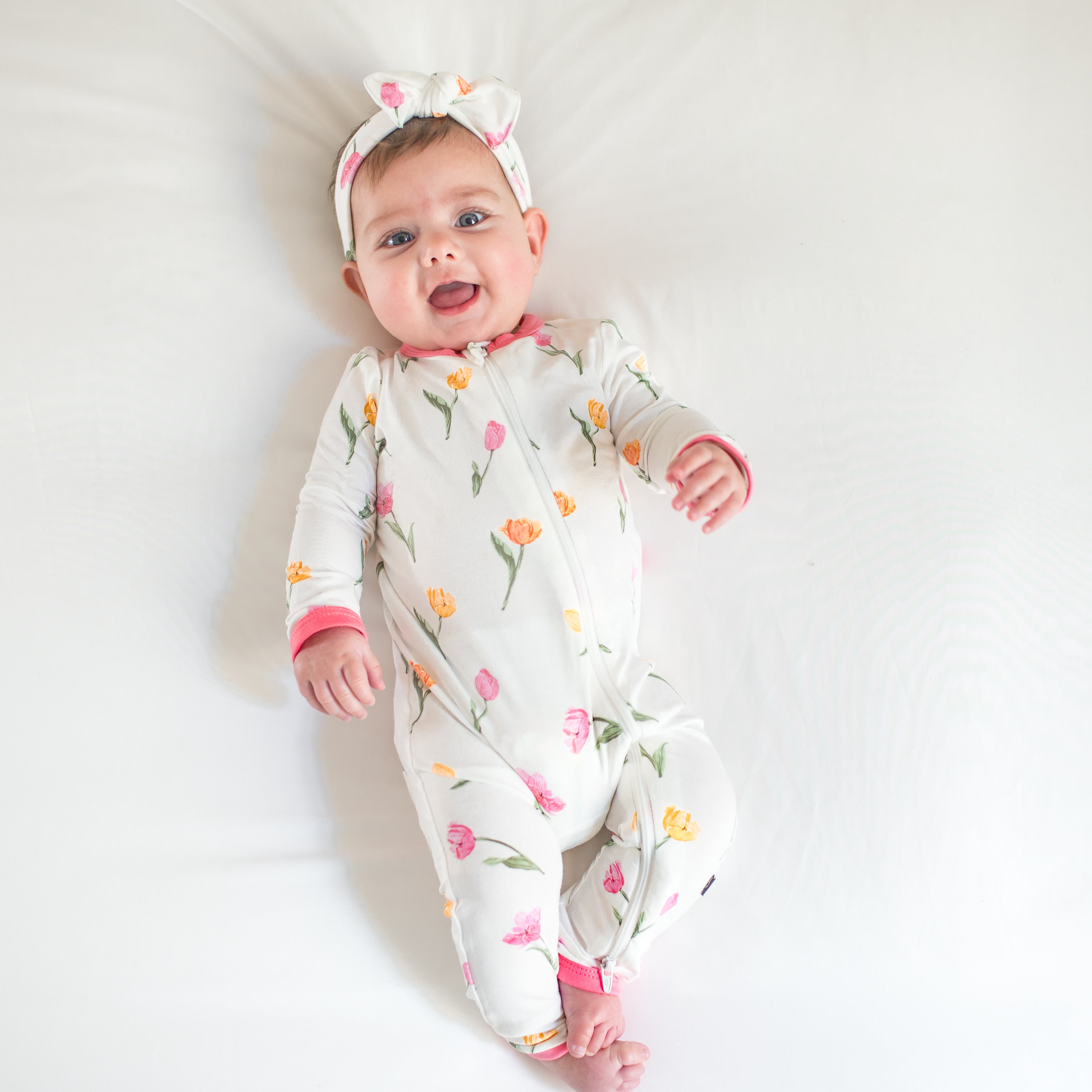 Smiling toddler laying on a bed wearing the Zippered Romper in Tulip with matching knotted bow headband